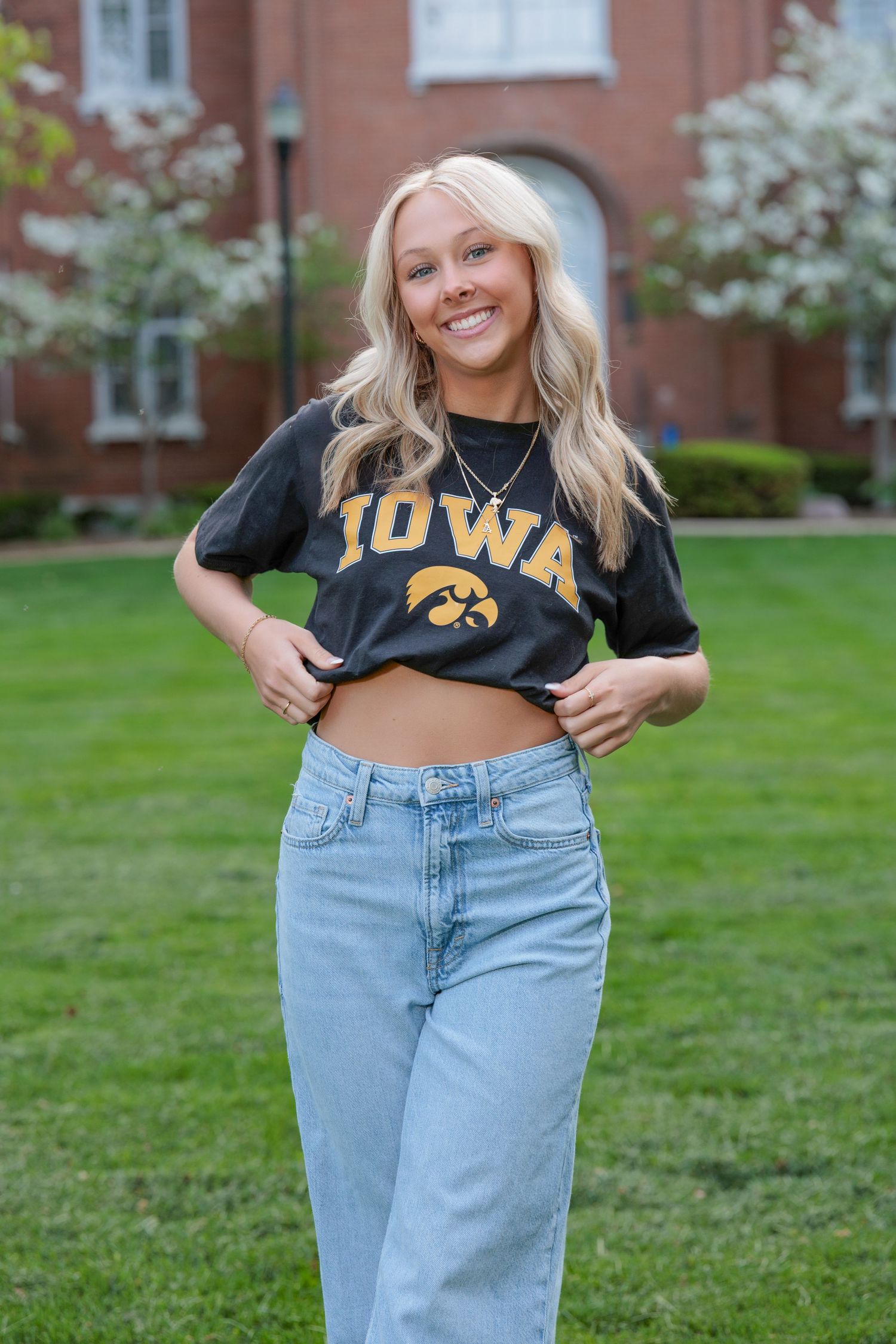 A young woman wearing a cropped black Iowa Hawkeyes t-shirt and light blue jeans poses on a grassy lawn.
