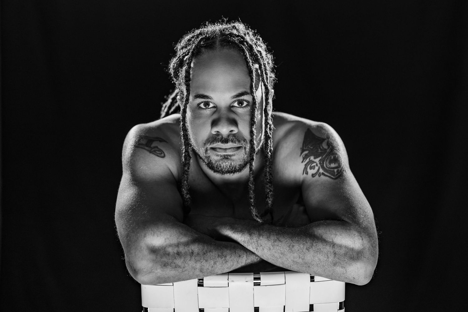 A dramatic black and white portrait of a muscular person with braids crossing their arms against their chest.