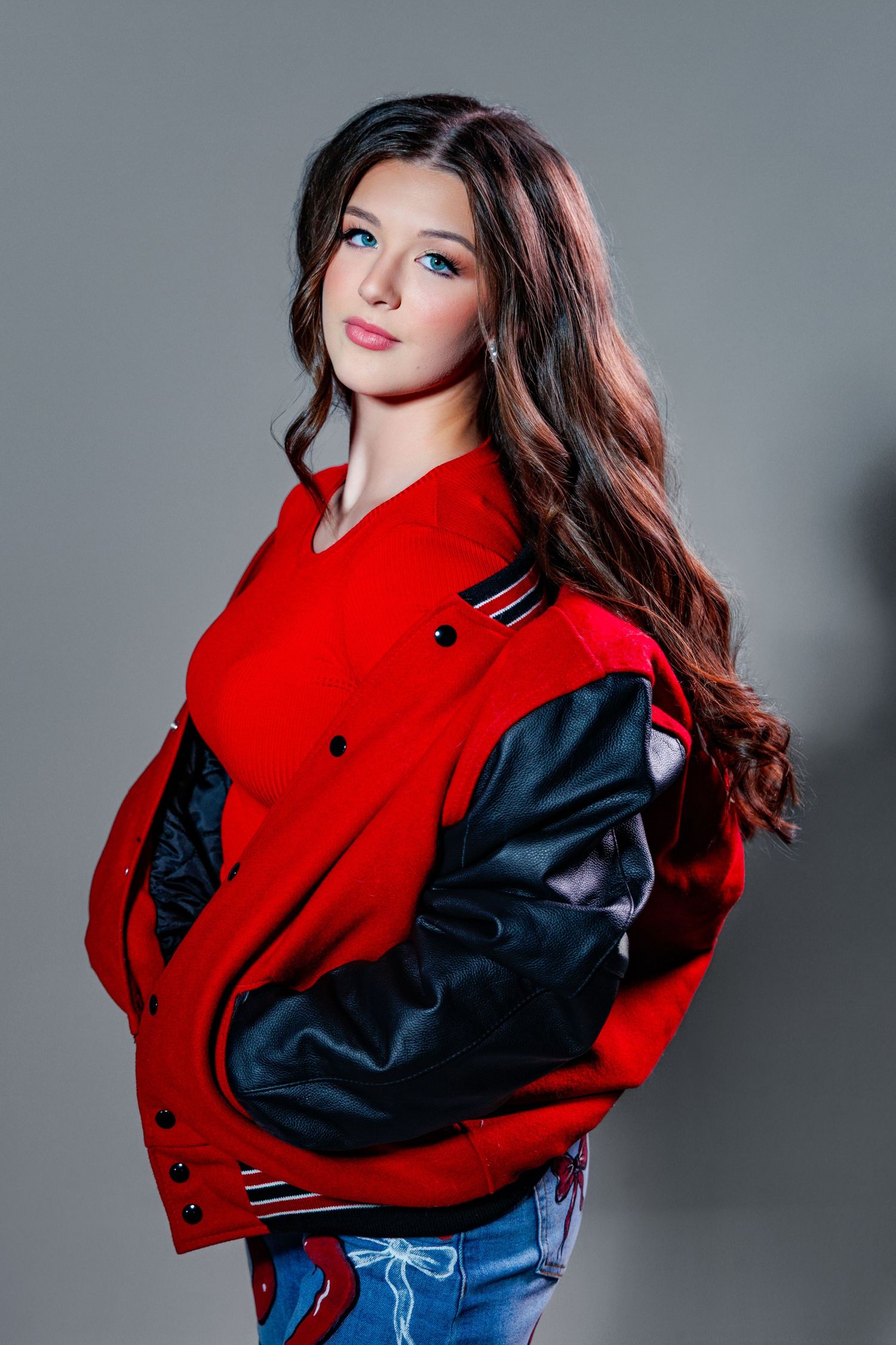Fashion model wearing red varsity jacket with black leather sleeves and blue jeans in studio photoshoot.