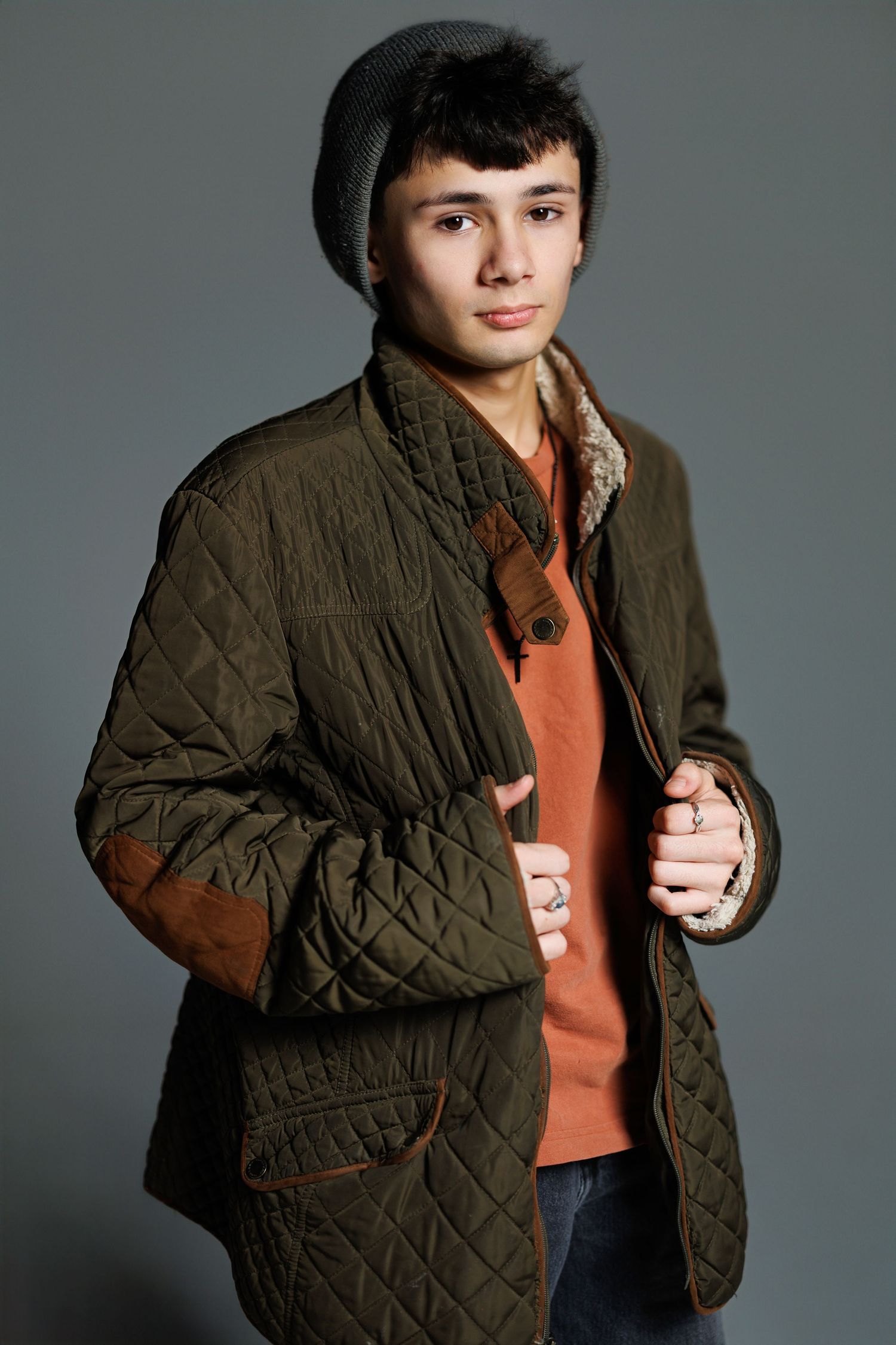 A person poses in a dark olive quilted jacket and orange sweater against a gray studio backdrop in multiple shots.
