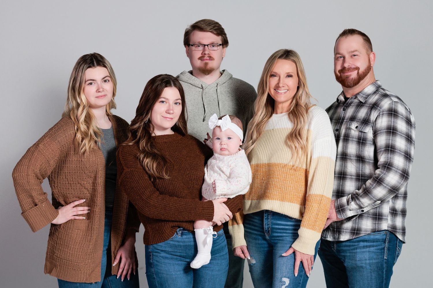 A multi-generational family group poses together in coordinating casual outfits of earth tones and plaid against a white backdrop.