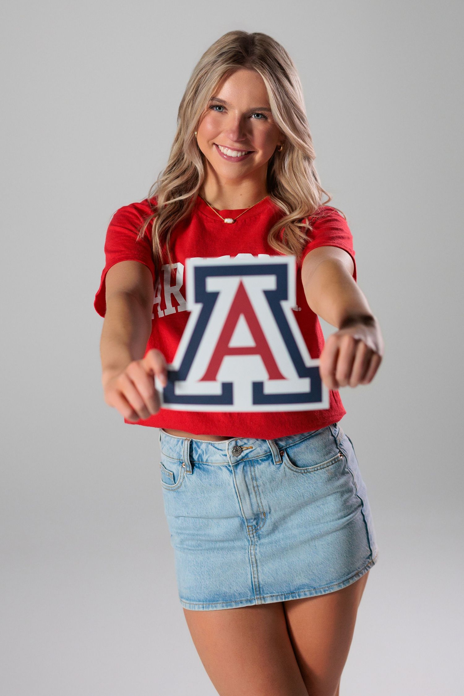 Person wearing a red University of Arizona t-shirt and denim mini skirt poses against a gray background.
