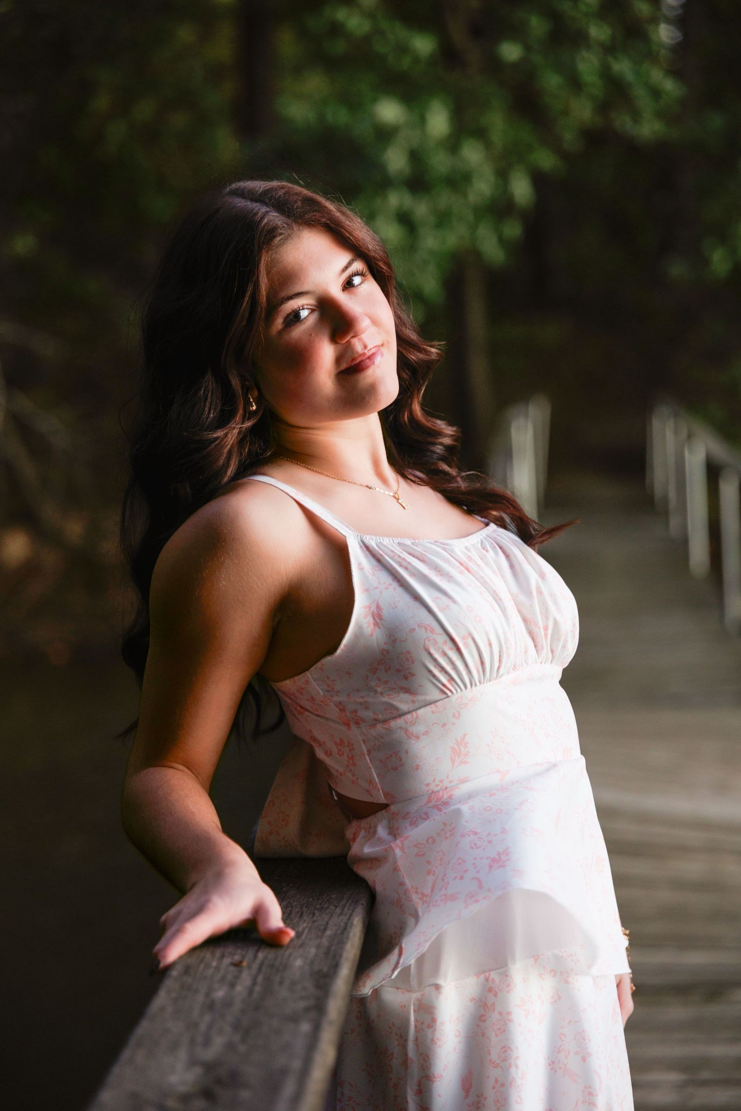 A person in a flowing white summer dress poses gracefully against a tree-lined path in natural outdoor lighting.