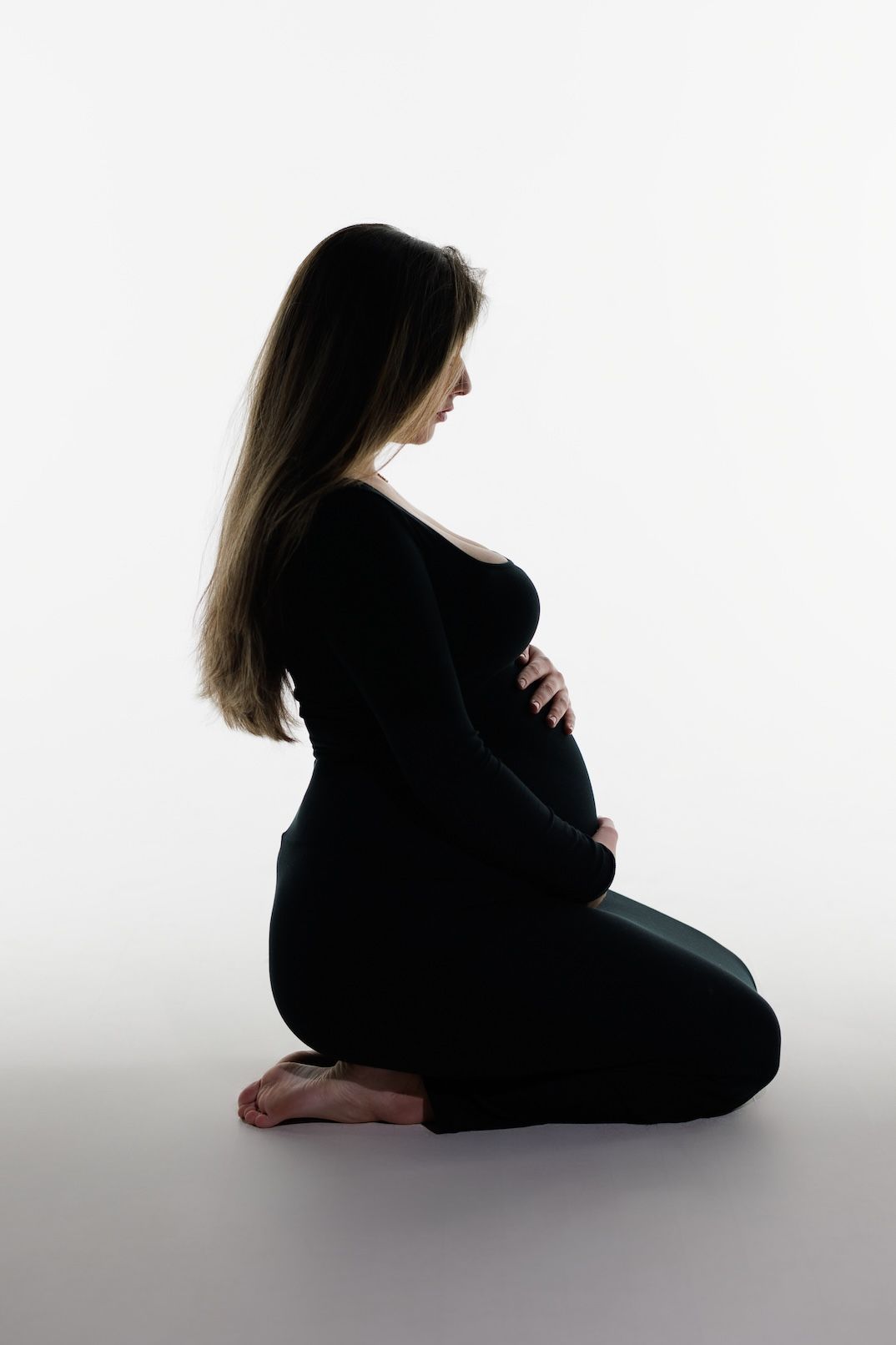 A silhouetted side view of a pregnant woman in black attire kneeling on white background.