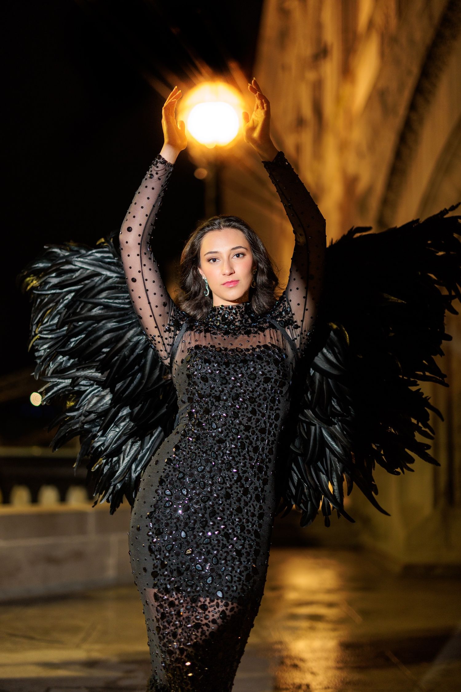 Figure in a sparkly black dress with feather wings holds a glowing orb above their head at night.