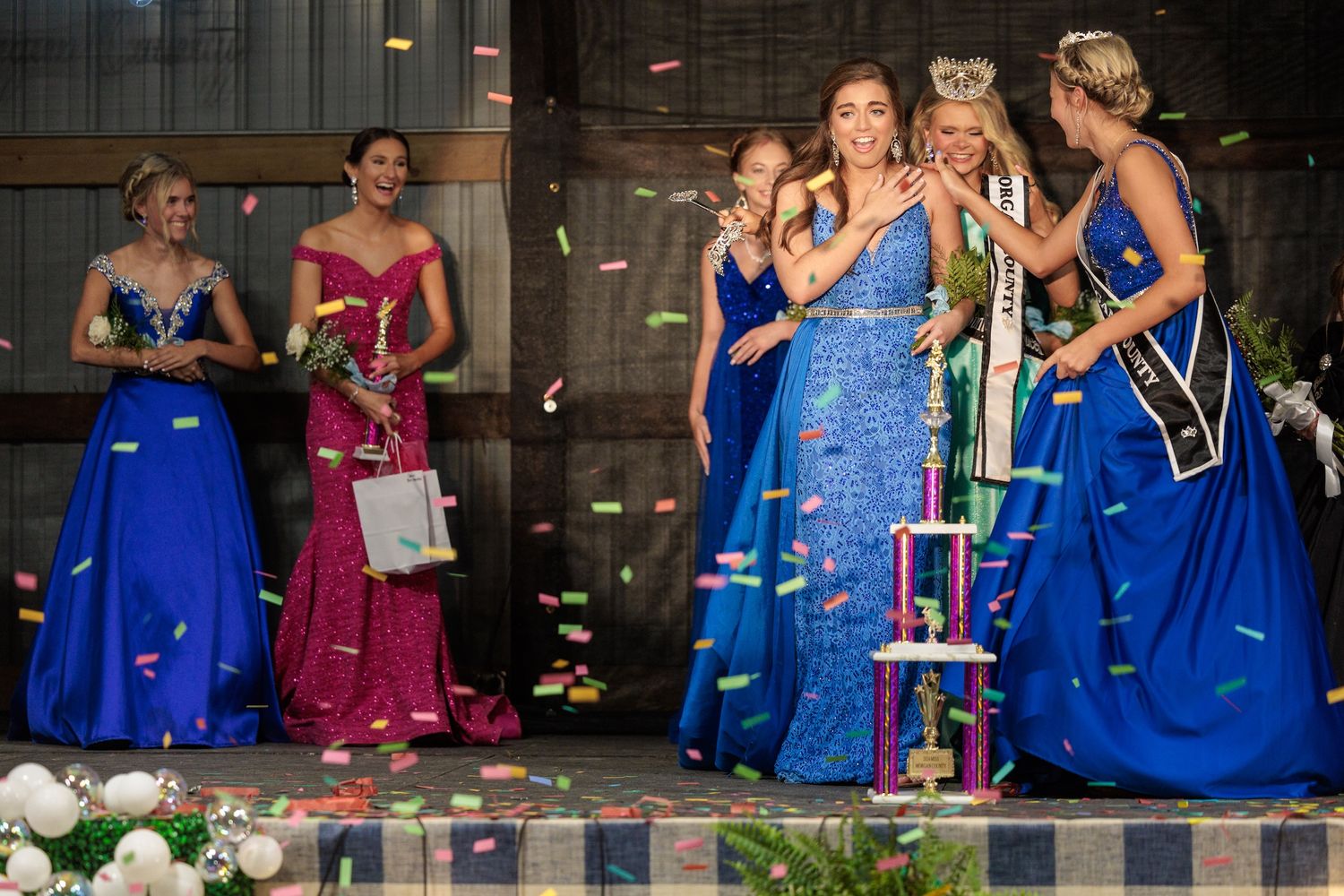 A group in bright blue and pink formal gowns gather around a trophy while confetti falls in an evening celebration.