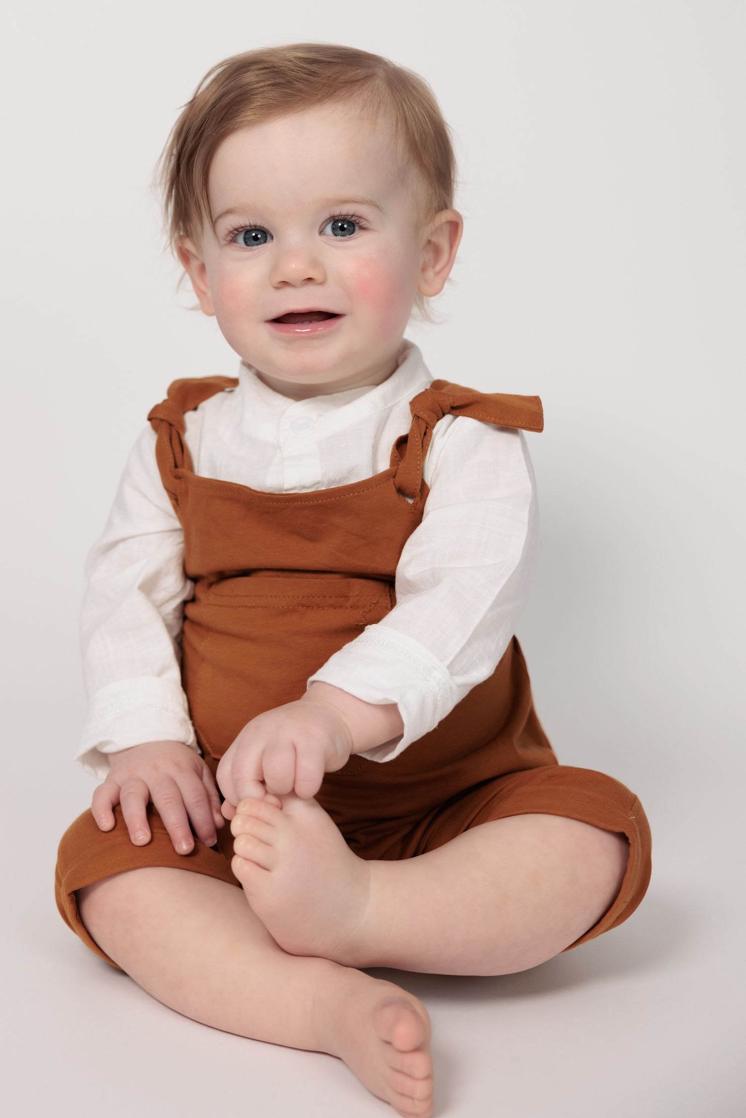 Baby wearing brown overalls and white long sleeve shirt sitting on light gray background poses sweetly.