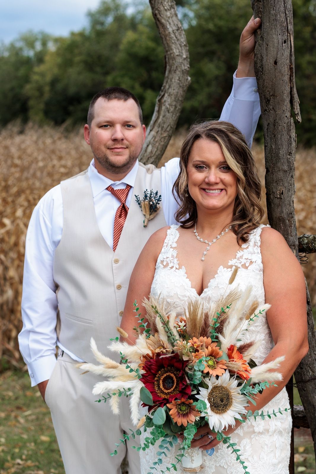 A couple poses outdoors on their wedding day with a beautiful bouquet featuring sunflowers and burgundy blooms.