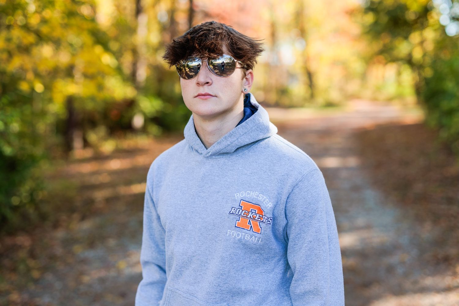 Someone wearing a light grey hoodie with orange logo and sunglasses poses on an autumn path with golden trees in background.