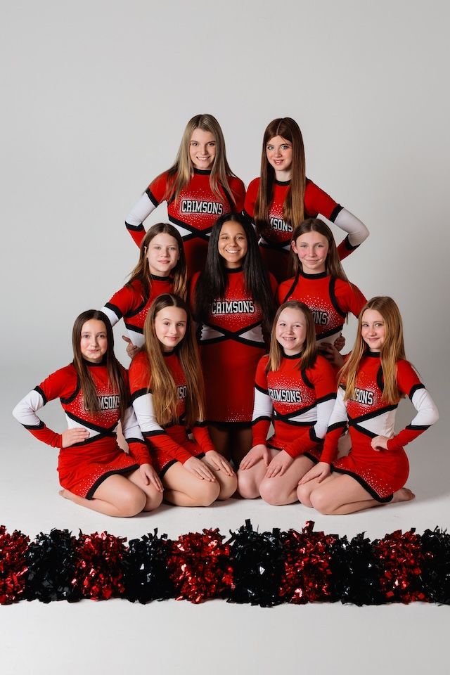 A cheerleading team poses in red and white uniforms with red decorative garland at their feet in a studio setting.