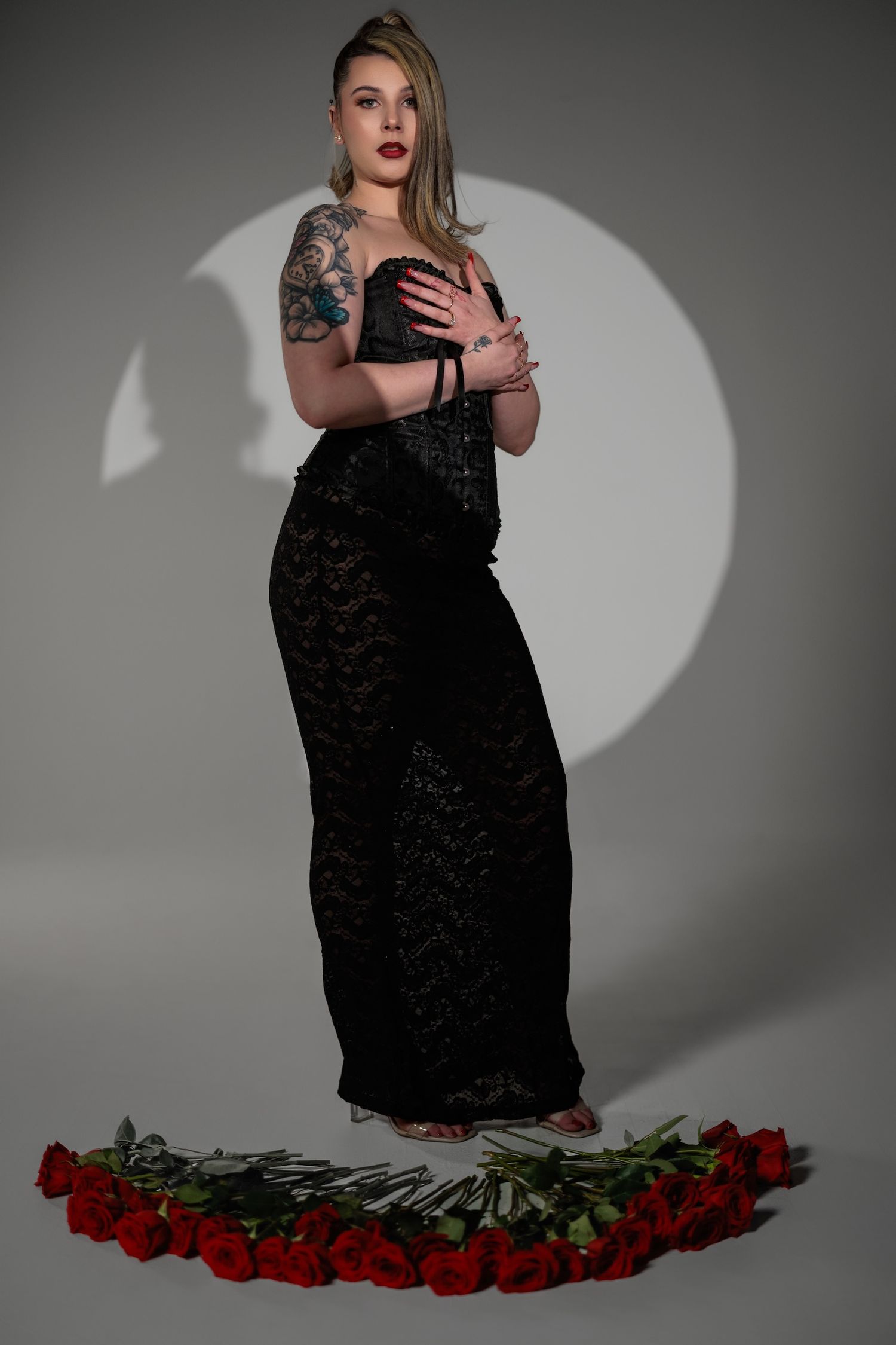 Artistic fashion portrait with figure in black lace dress standing above circle of red roses against gray backdrop.