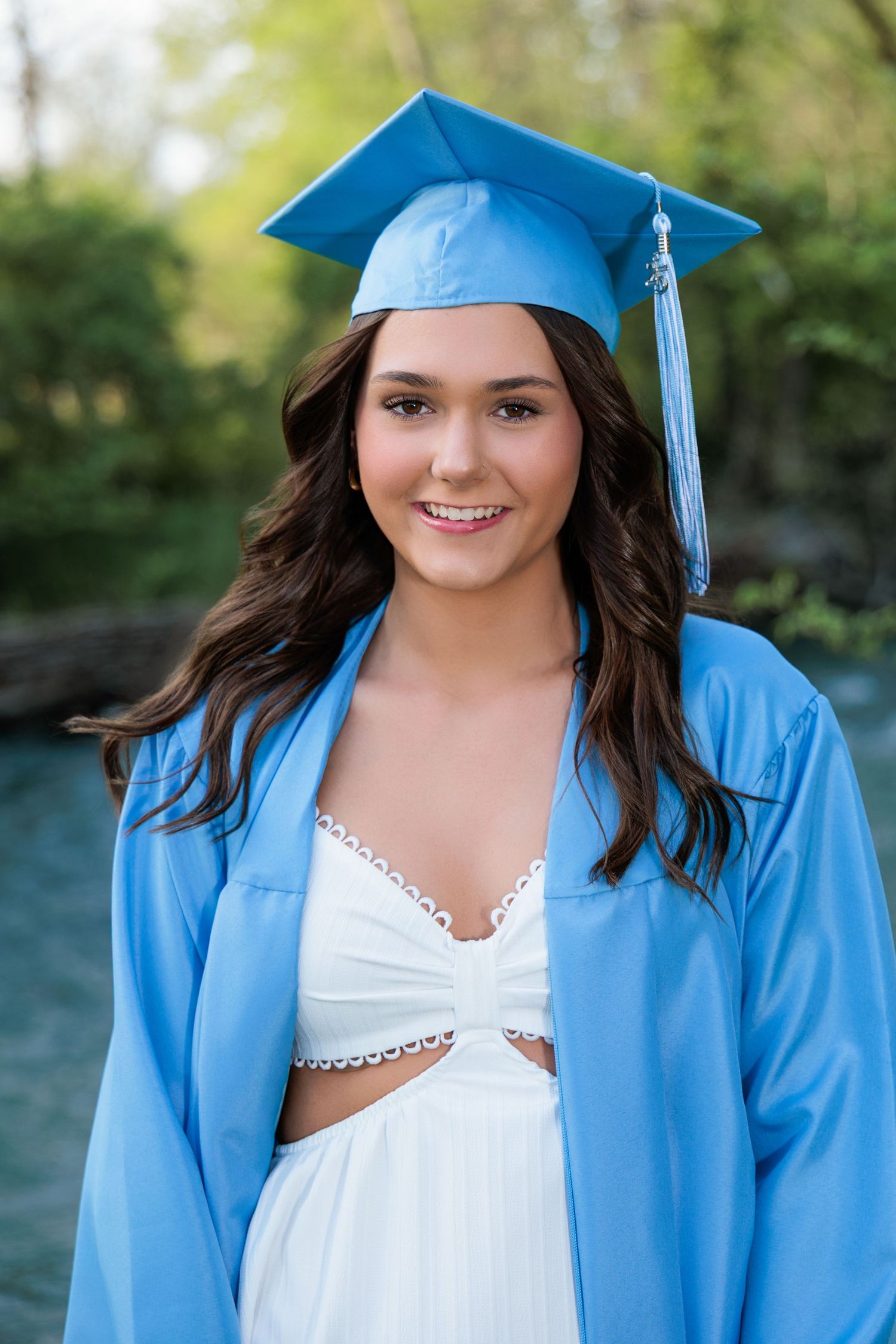 Graduate wearing light blue cap and gown with white dress smiles while posing for outdoor graduation photo.