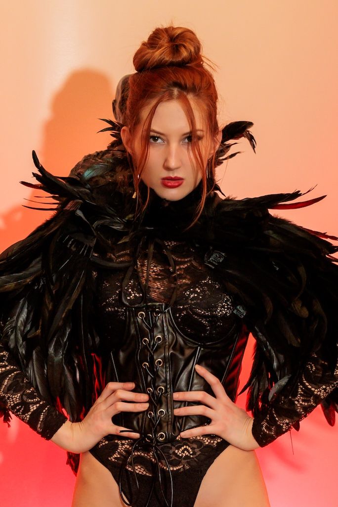 Dark feathered bodysuit with corset detail against pink and orange gradient background.