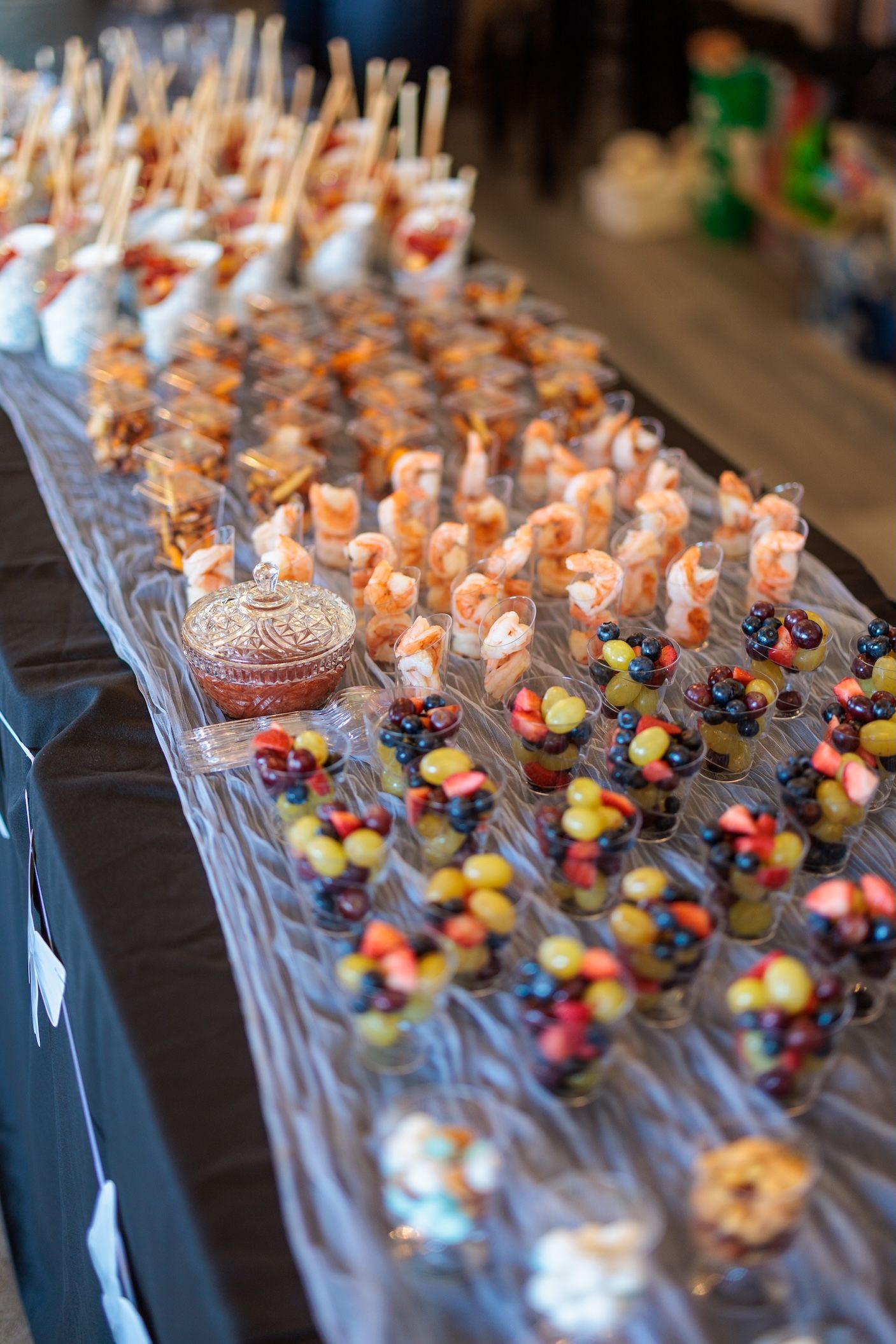 A catering display table with rows of appetizers including shrimp cocktails and colorful fruit cups arranged on a black tablecloth.