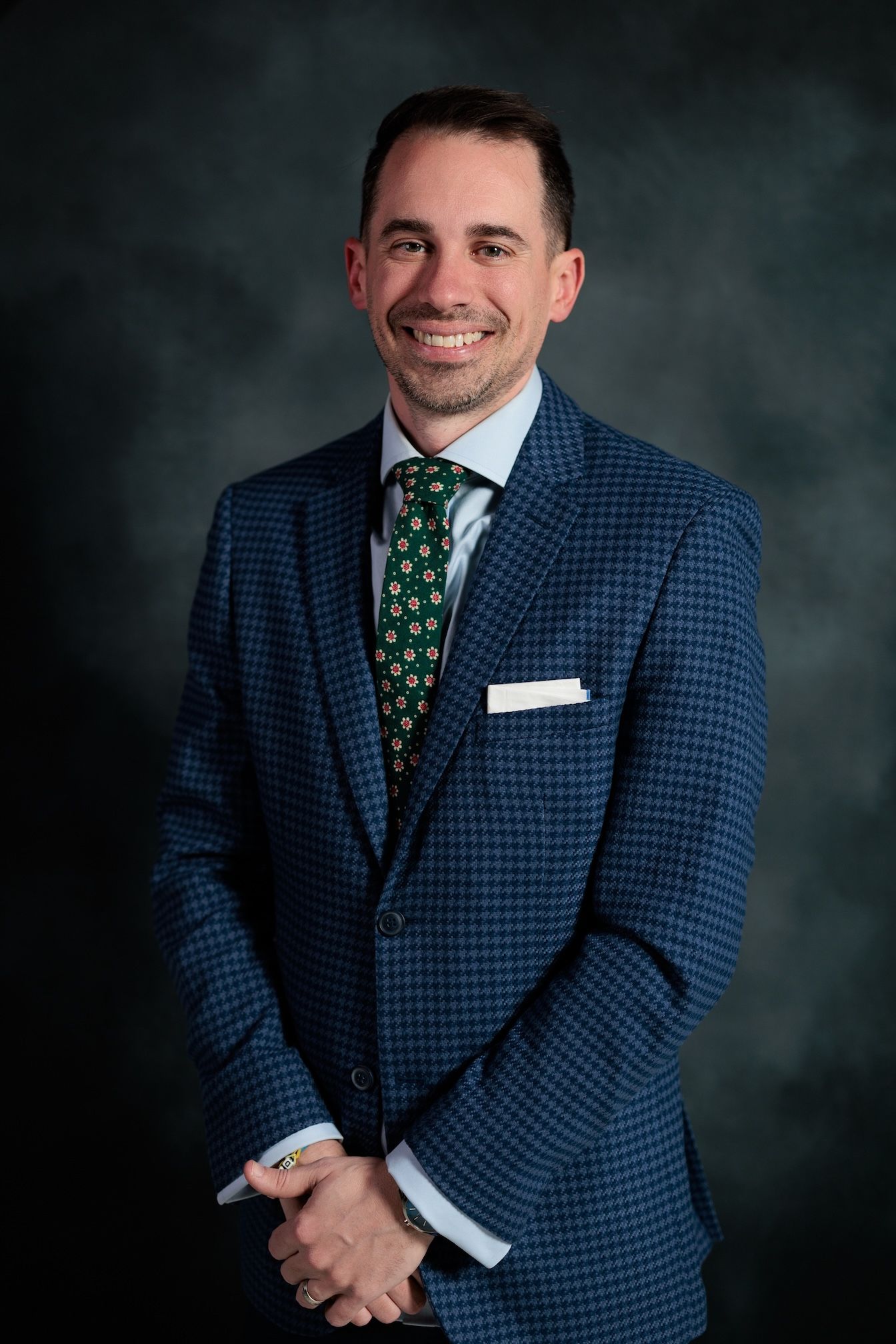 Professional headshot of person in navy checkered suit and green patterned tie against dark background.