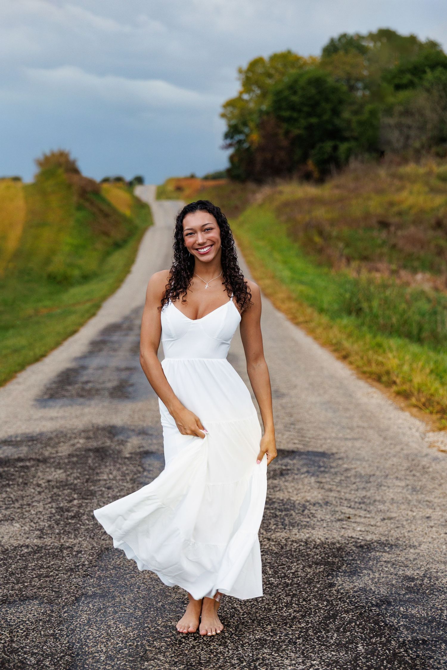 A white flowing sundress moves in the breeze on a rural country road surrounded by green hills on a cloudy day.