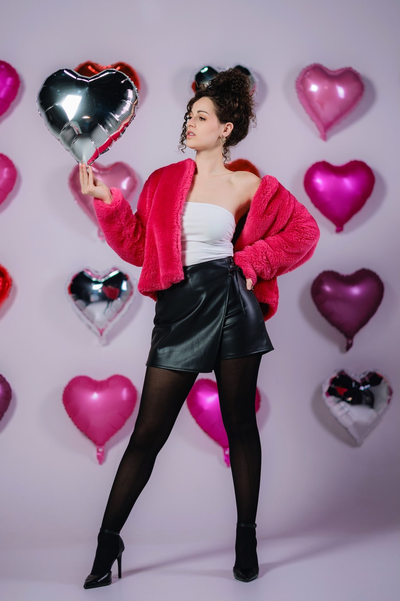 Fashion model poses against heart balloon backdrop wearing red puffer jacket, white top and black skirt for Valentine's theme shoot.