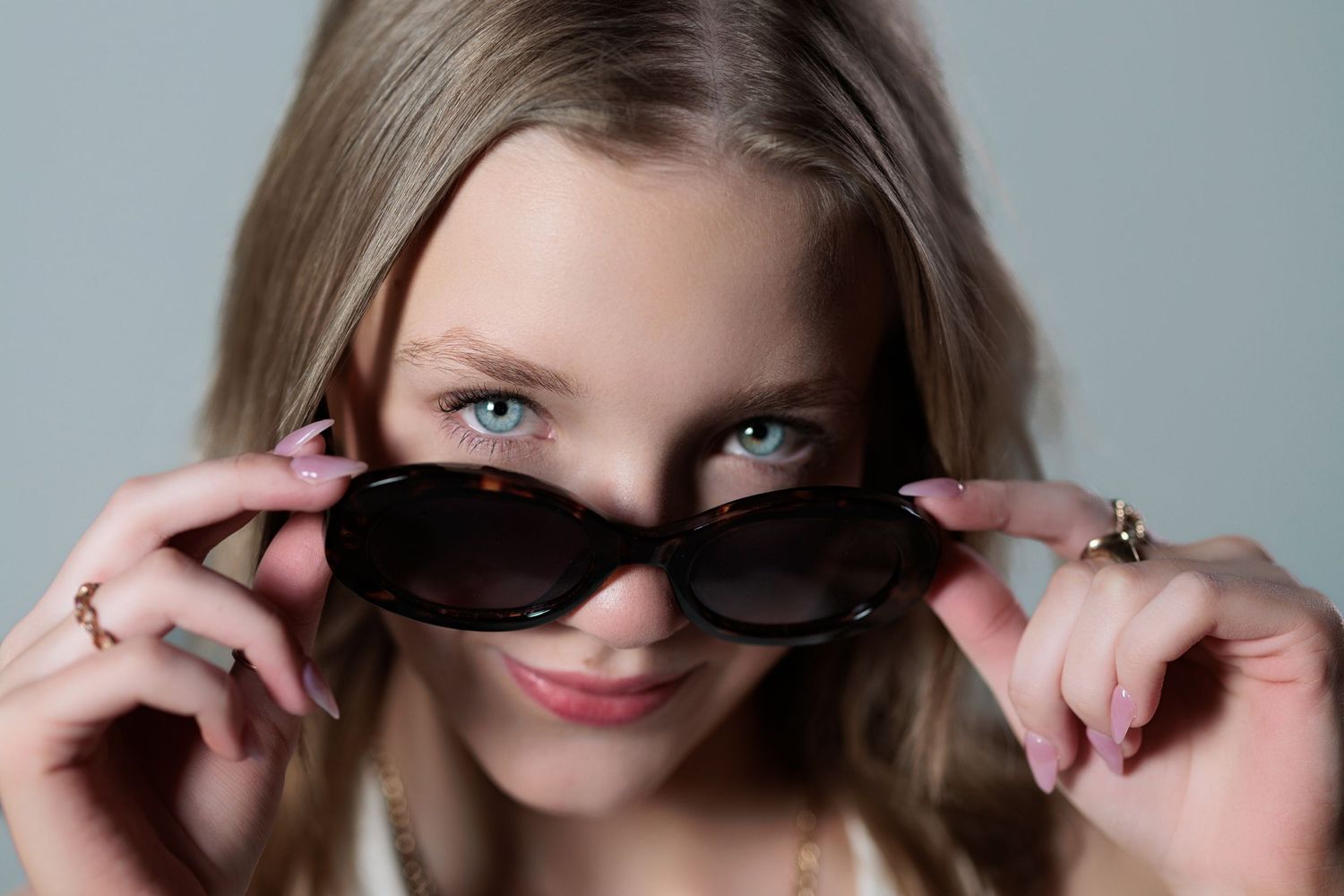 Looking over dark sunglasses with striking blue eyes, wearing rings on fingers holding the frames.