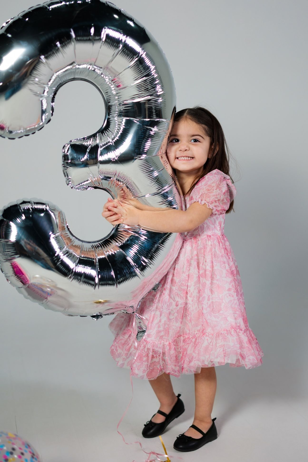 A child in a pink party dress poses with a large silver number 3 balloon against a gray background.