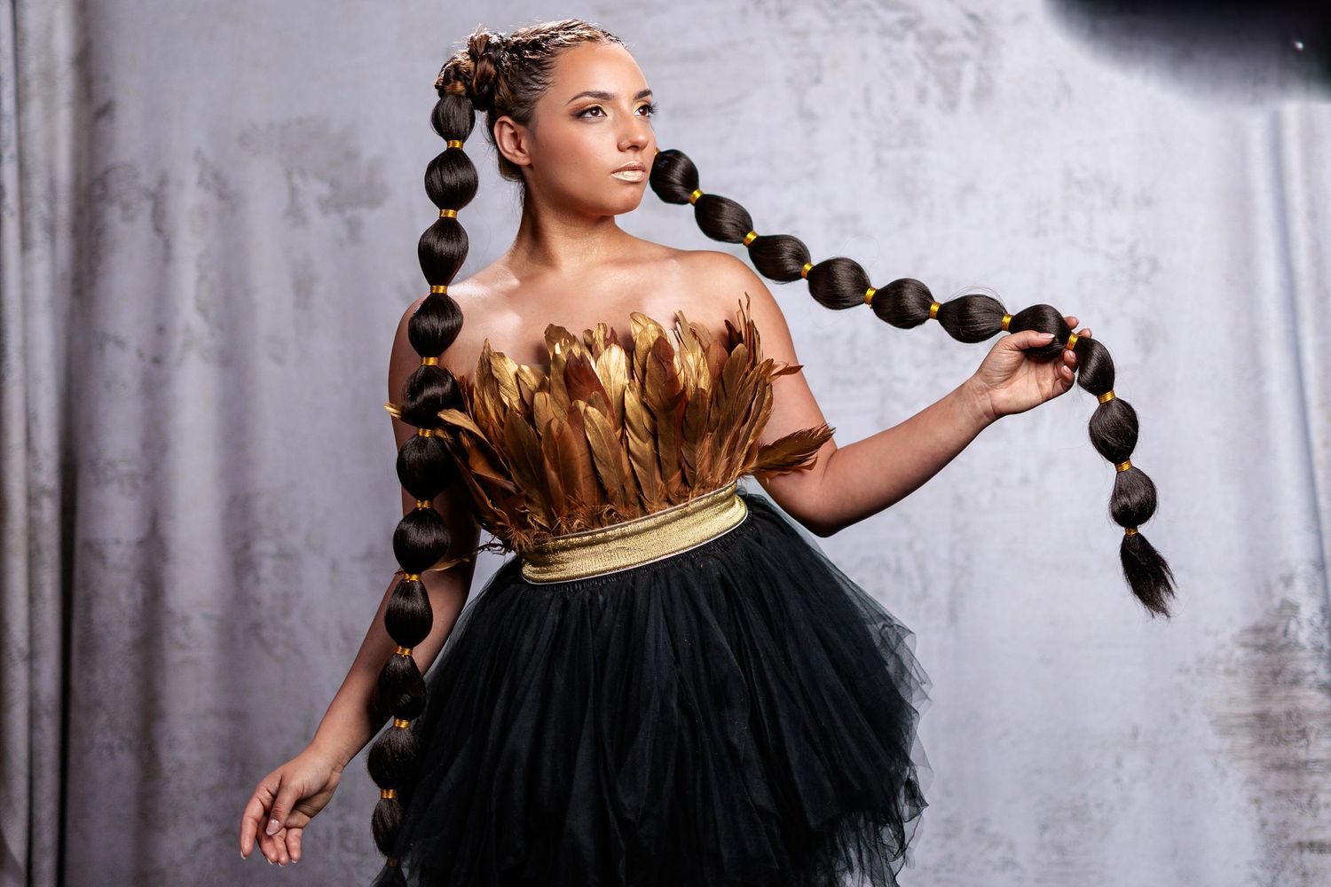 An elegant model wearing a metallic gold top and black tulle skirt poses with uniquely styled long braided hair.
