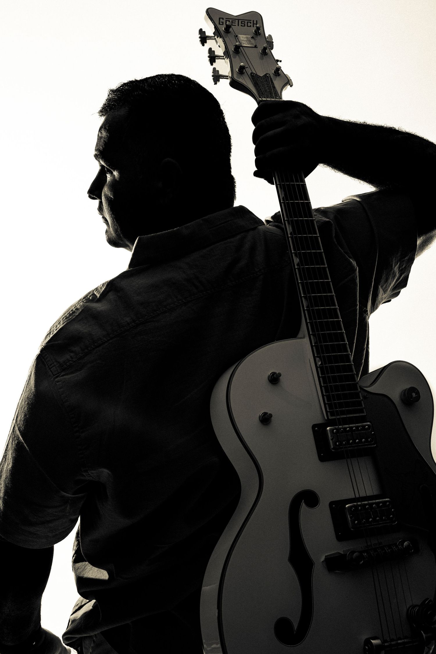 Silhouetted musician in black clothing holds an acoustic guitar in dramatic profile against white background.