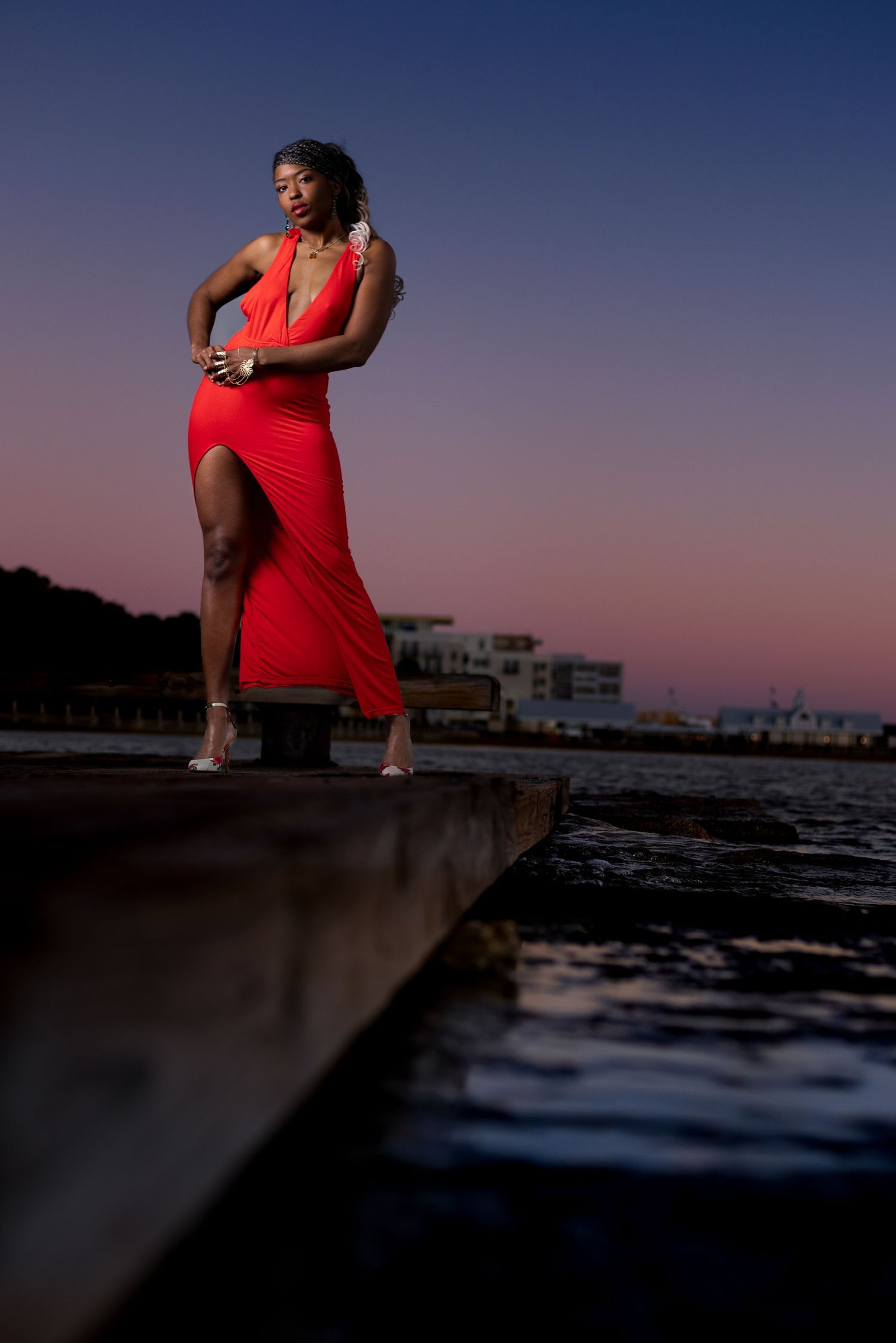 Figure in a striking red dress poses dramatically at sunset on a waterfront pier. Portrava Creative Studios