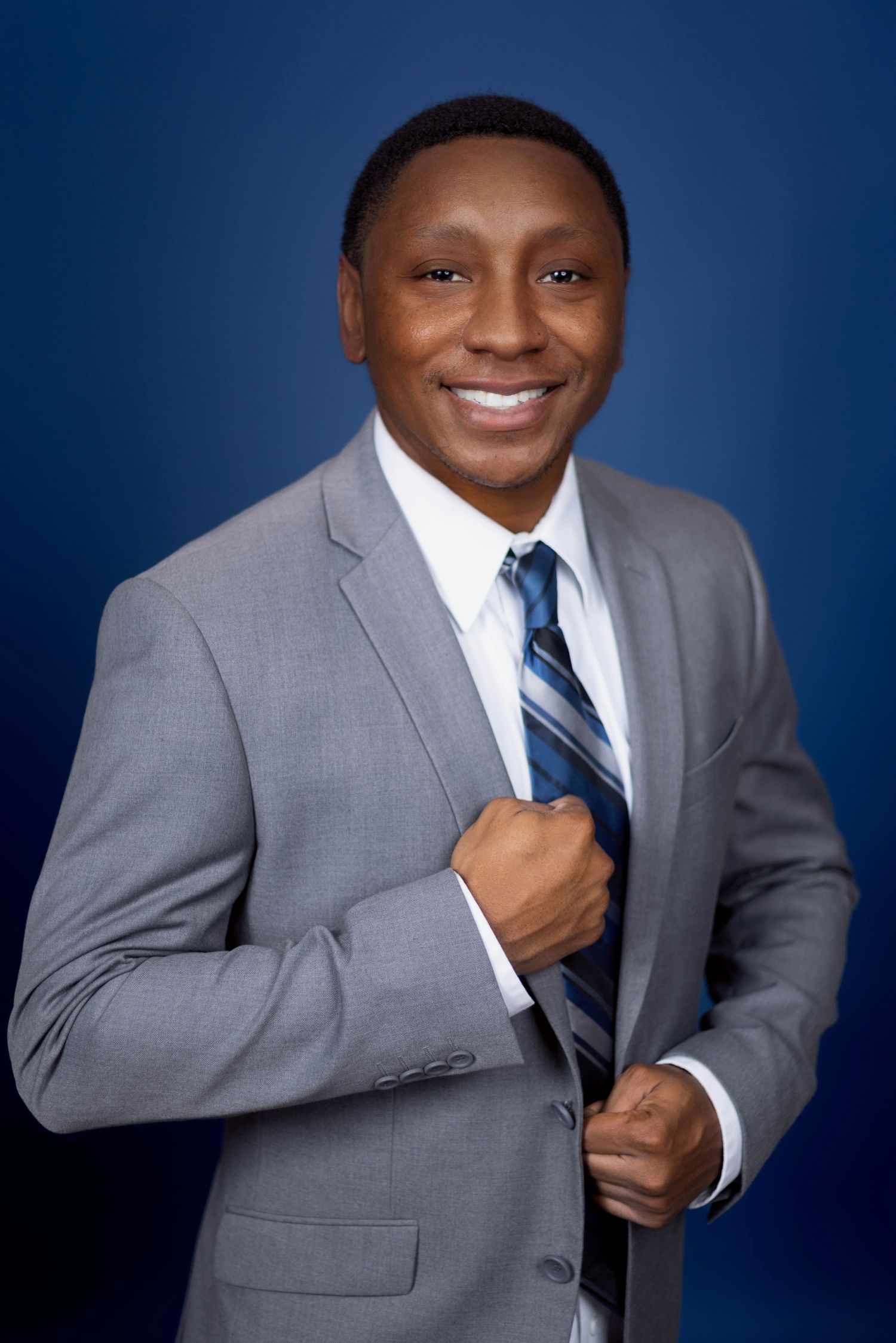 Professional portrait of someone in grey suit and striped tie smiling against blue background. Portrava Creative Studios