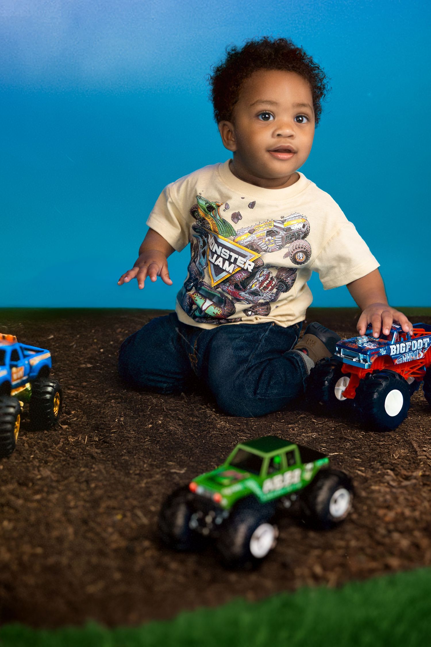 Young child playing with colorful toy monster trucks on dirt and grass surface against blue background. Portrava Creative Studios