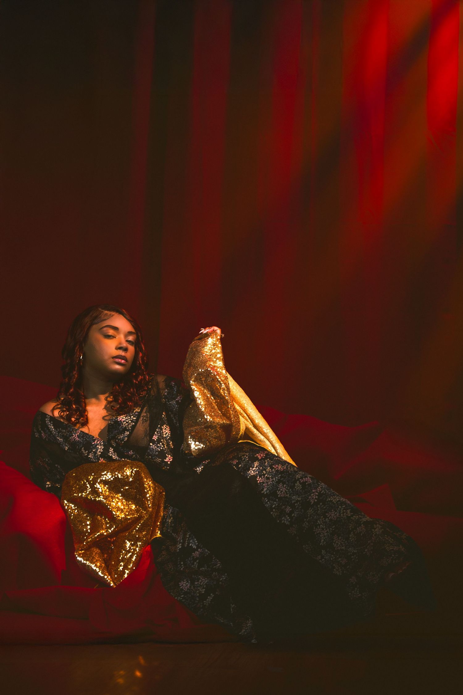 Model wearing black and gold ensemble reclines elegantly in moody red lighting. Portrava Creative Studios