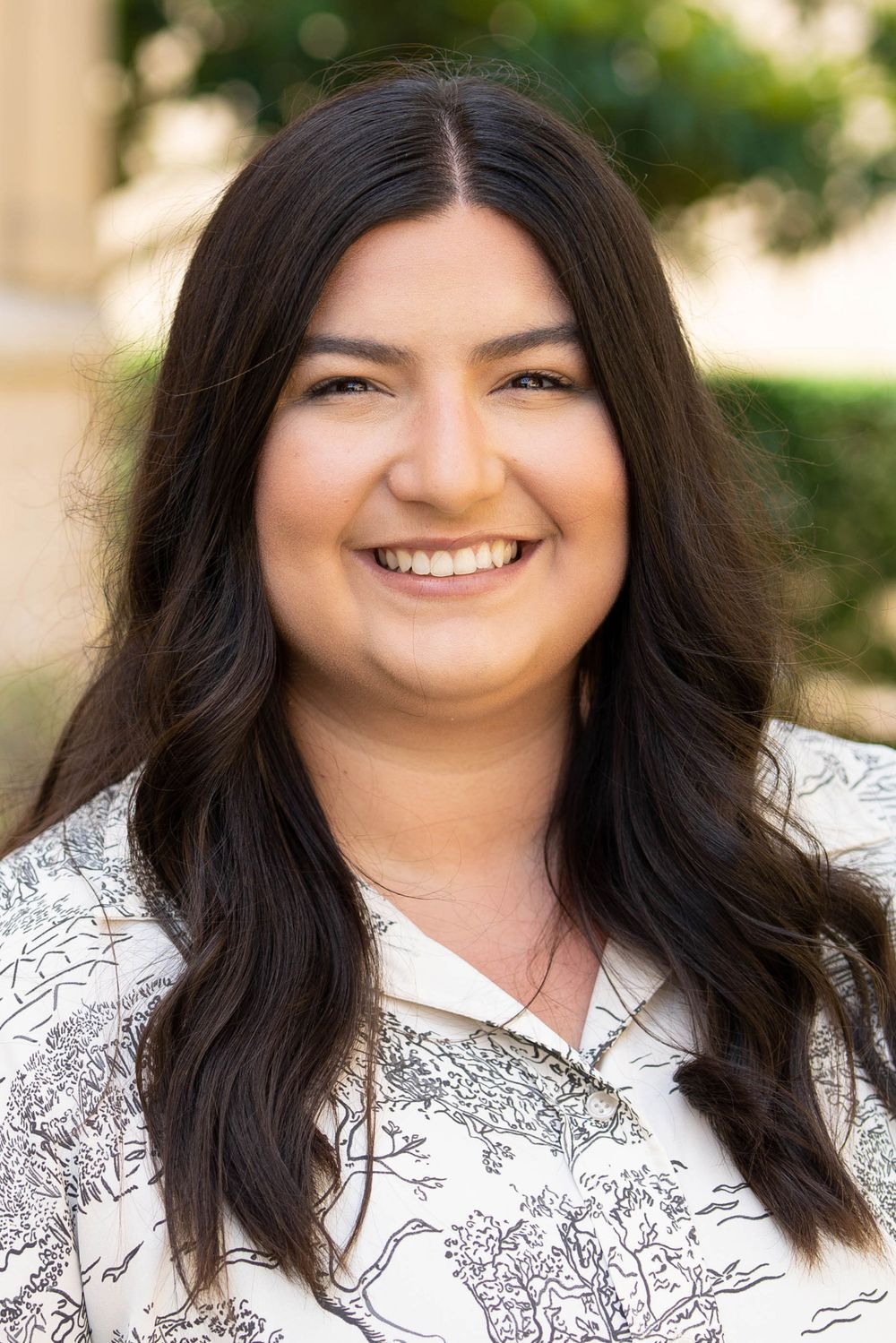 Professional headshot of person with long dark hair wearing floral patterned white top. Portrava Creative Studios