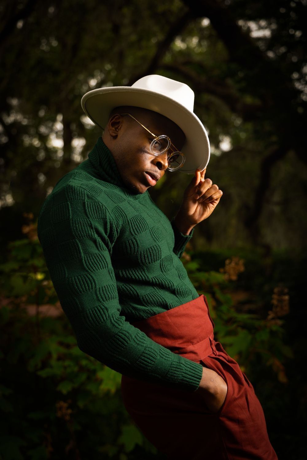Stylish person in green sweater and white hat poses outdoors in natural setting. Portrava Creative Studios