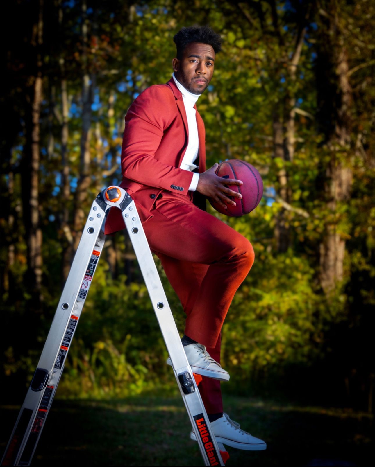 Person in red suit poses with basketball on white ladder outdoors. Portrava Creative Studios