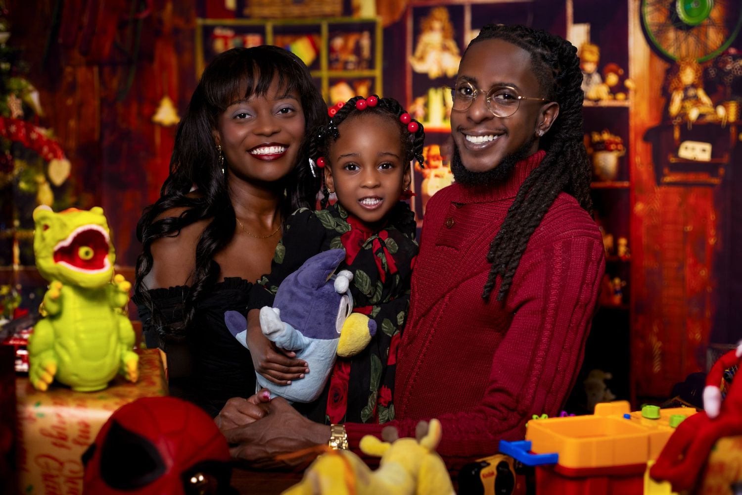 A family smiles together while posing at a festive holiday setting with decorative toys and string lights in the background. Portrava