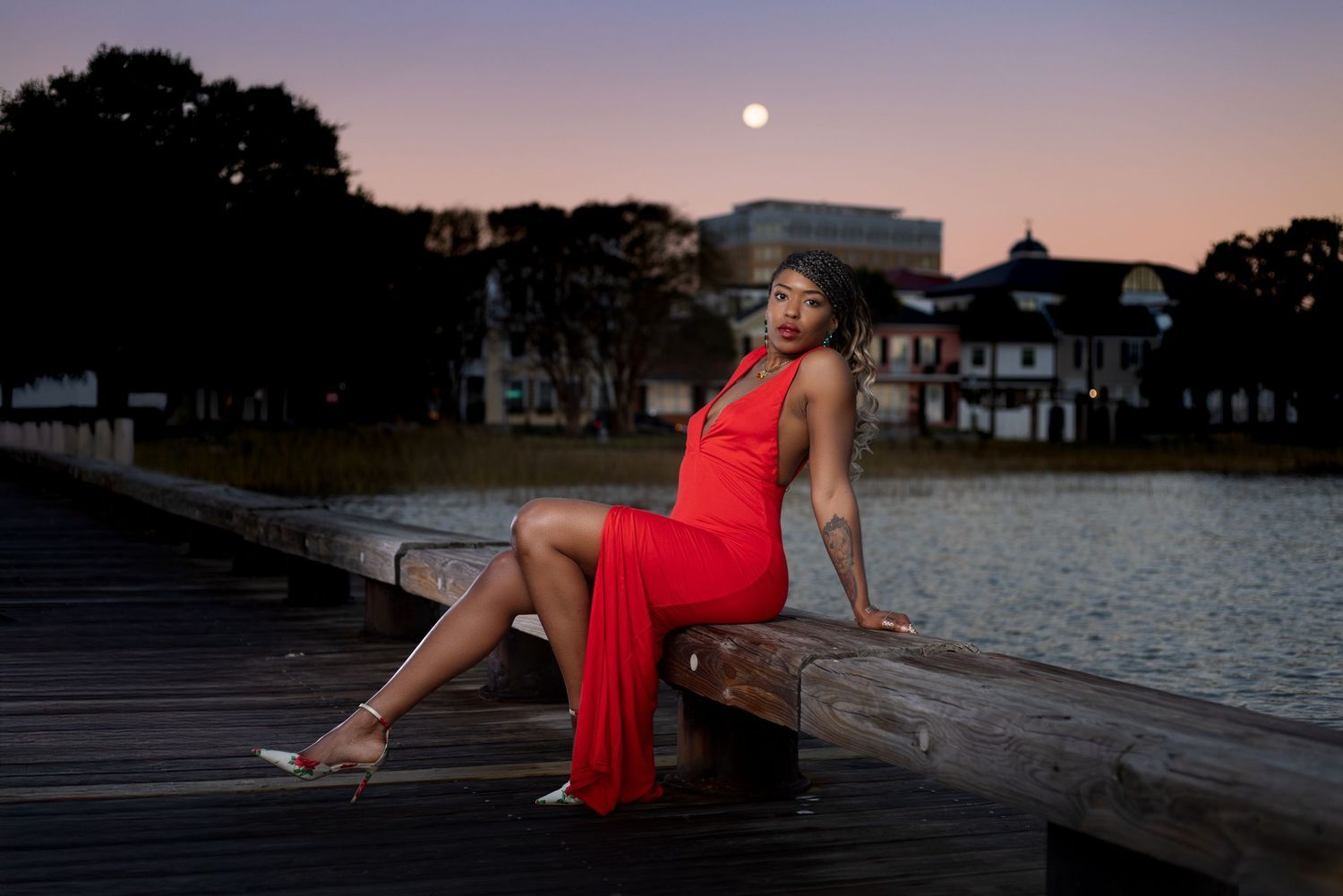 Sunset portrait of person in red dress reclining on wooden dock with moon rising. Portrava Creative Studios