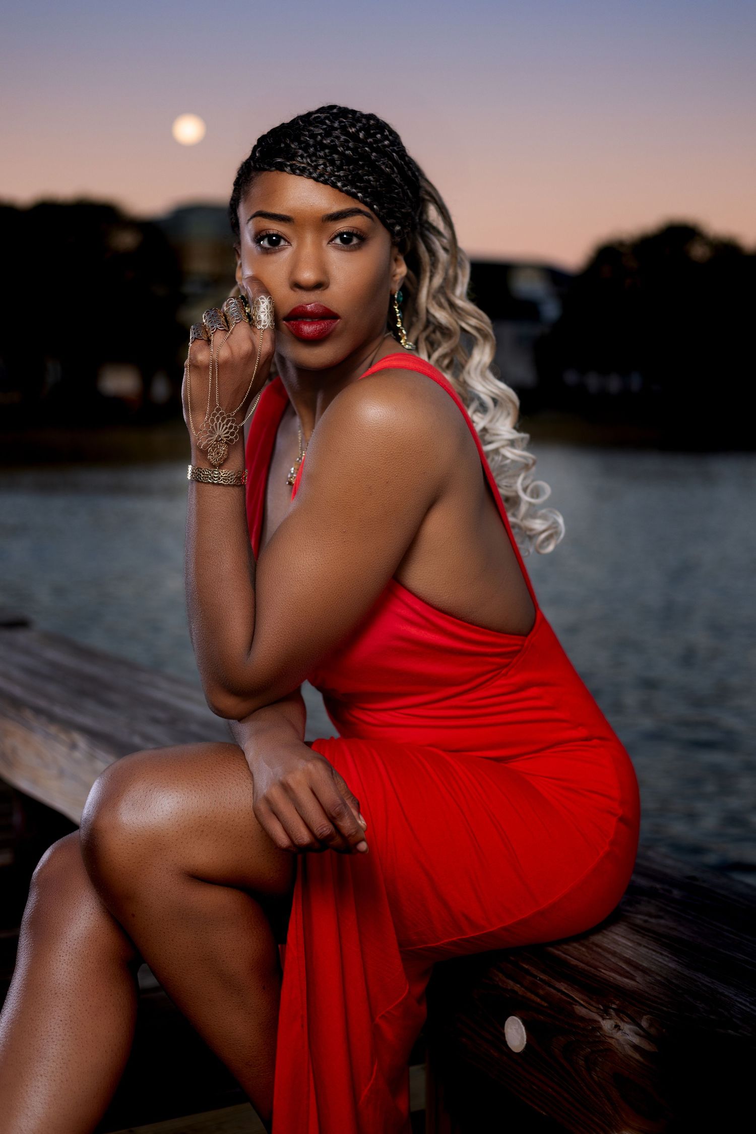 Woman in vibrant red dress poses elegantly against sunset waterfront backdrop. Portrava Creative Studios