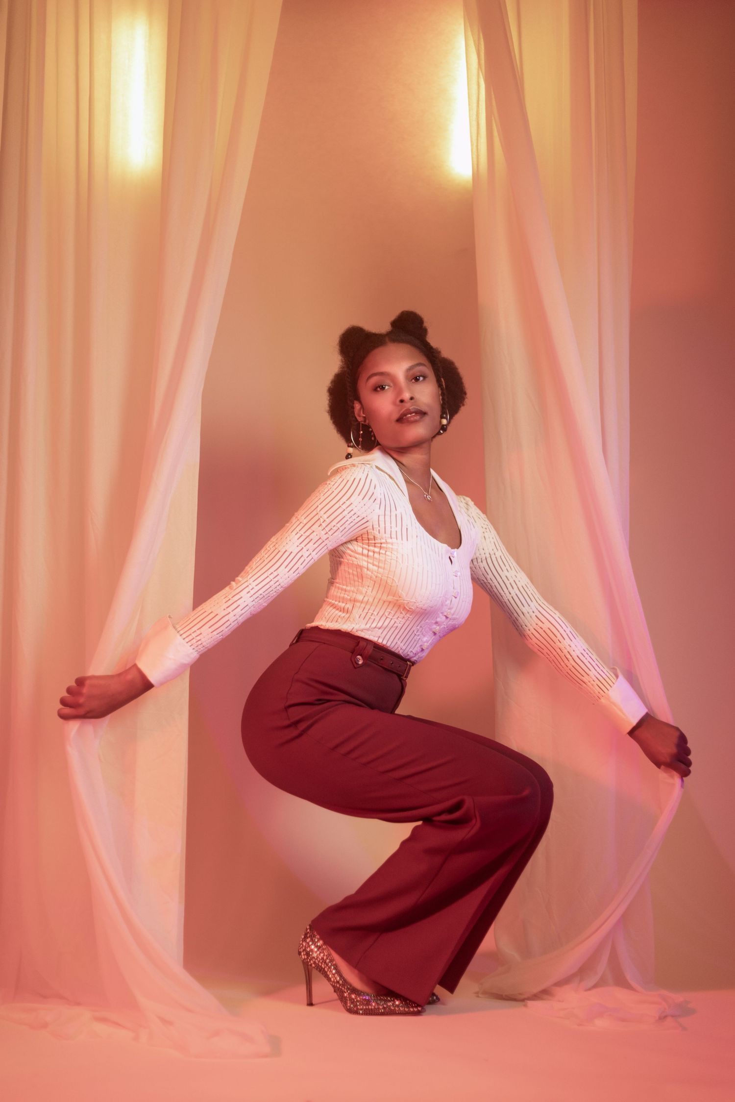 Elegant pose between flowing curtains wearing white top and burgundy pants in warm studio lighting. Portrava Creative Studios