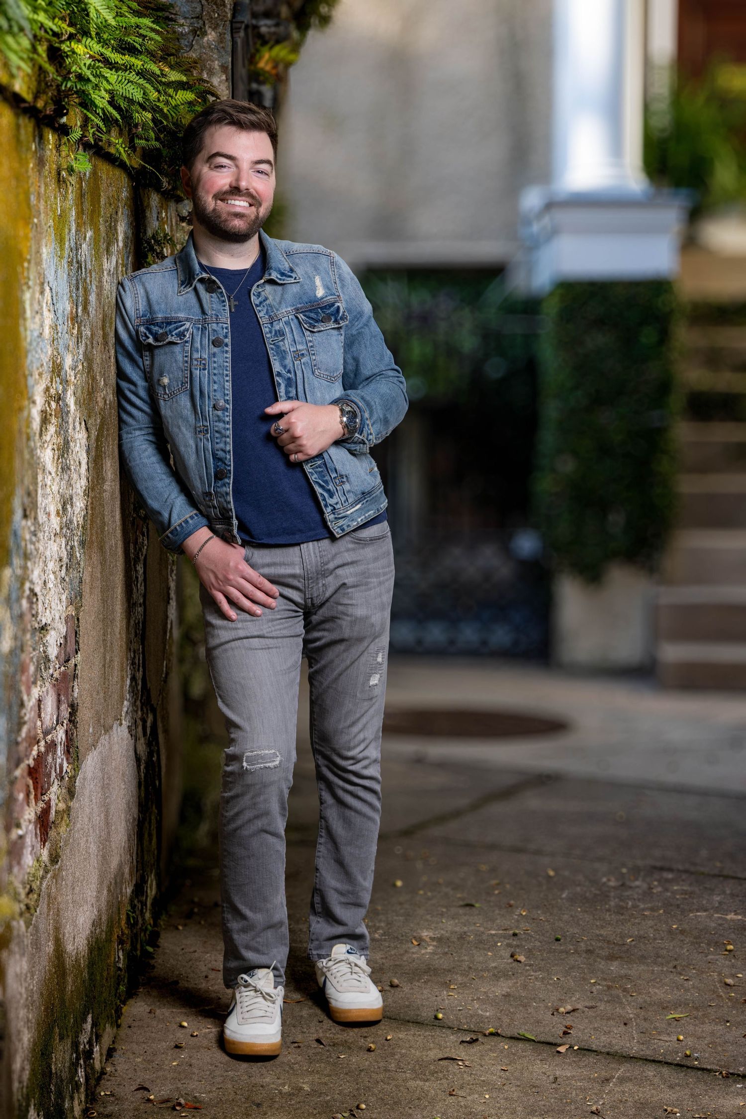 Casual portrait of person in denim jacket leaning against mossy wall with urban background. Portrava Creative Studios