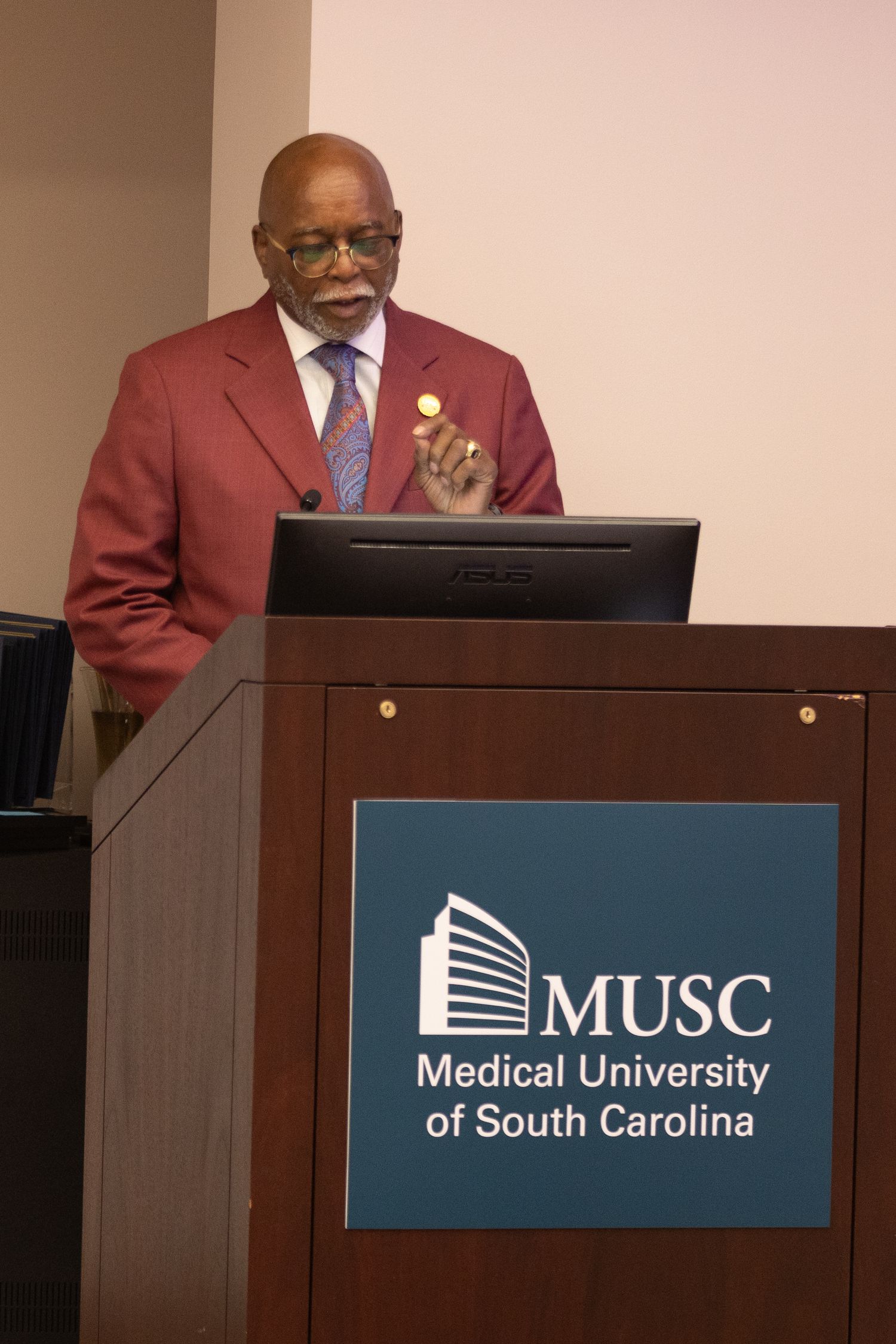 Speaker at podium delivering presentation at Medical University of South Carolina with MUSC logo displayed. Portrava Creative Studios