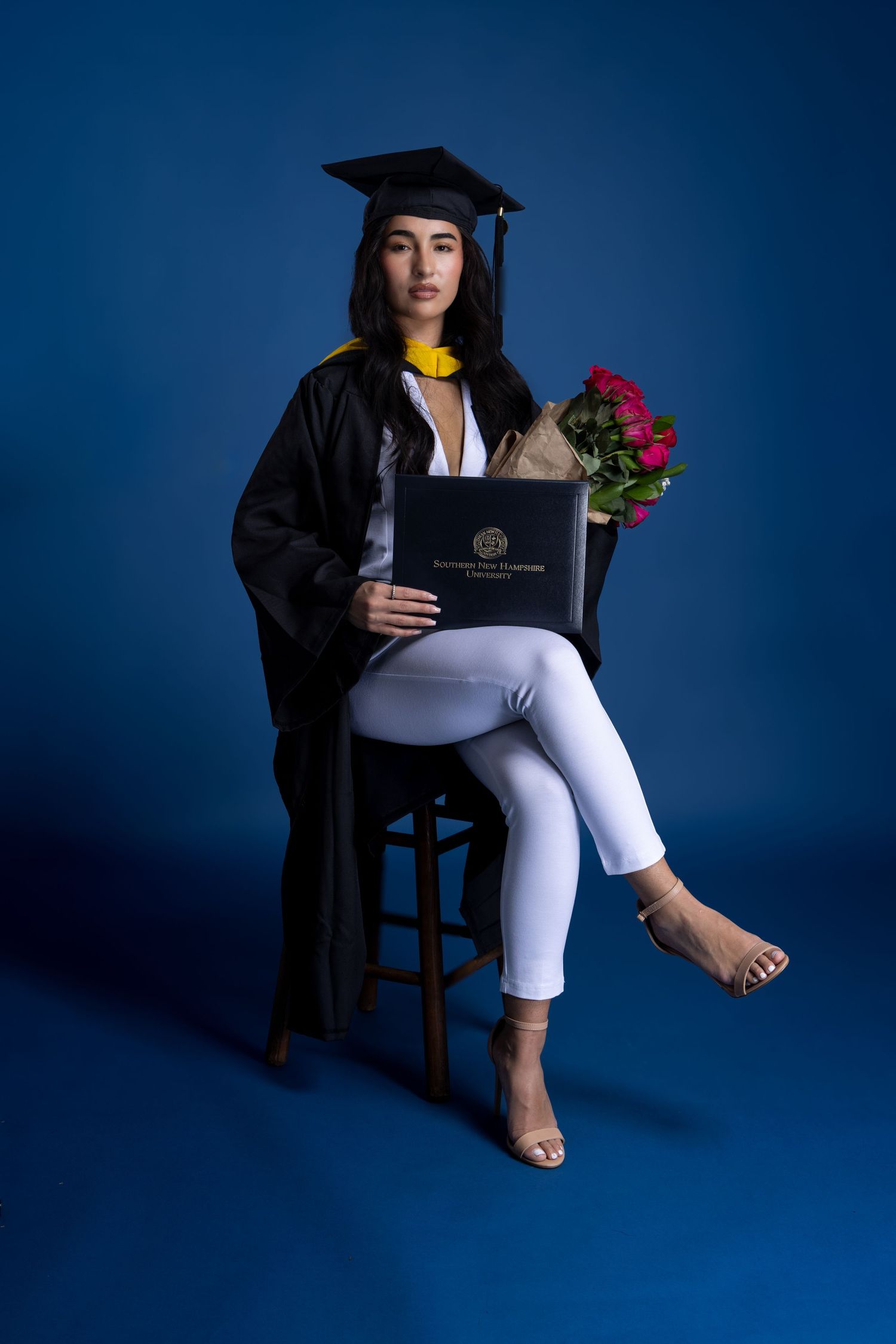 Graduate in cap and gown holds diploma and roses while seated on stool. Portrava Creative Studios