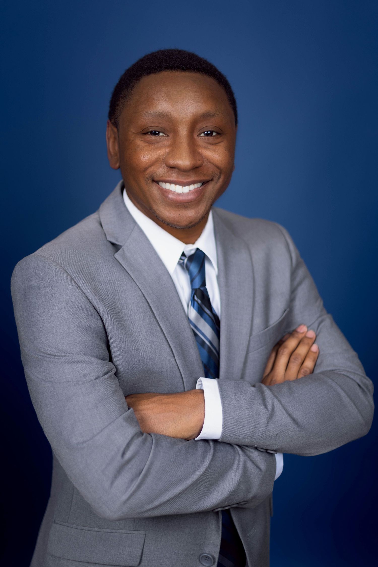 Professional headshot of a person wearing a gray suit and striped tie smiling warmly against a navy blue background.Portrava Creative Studio