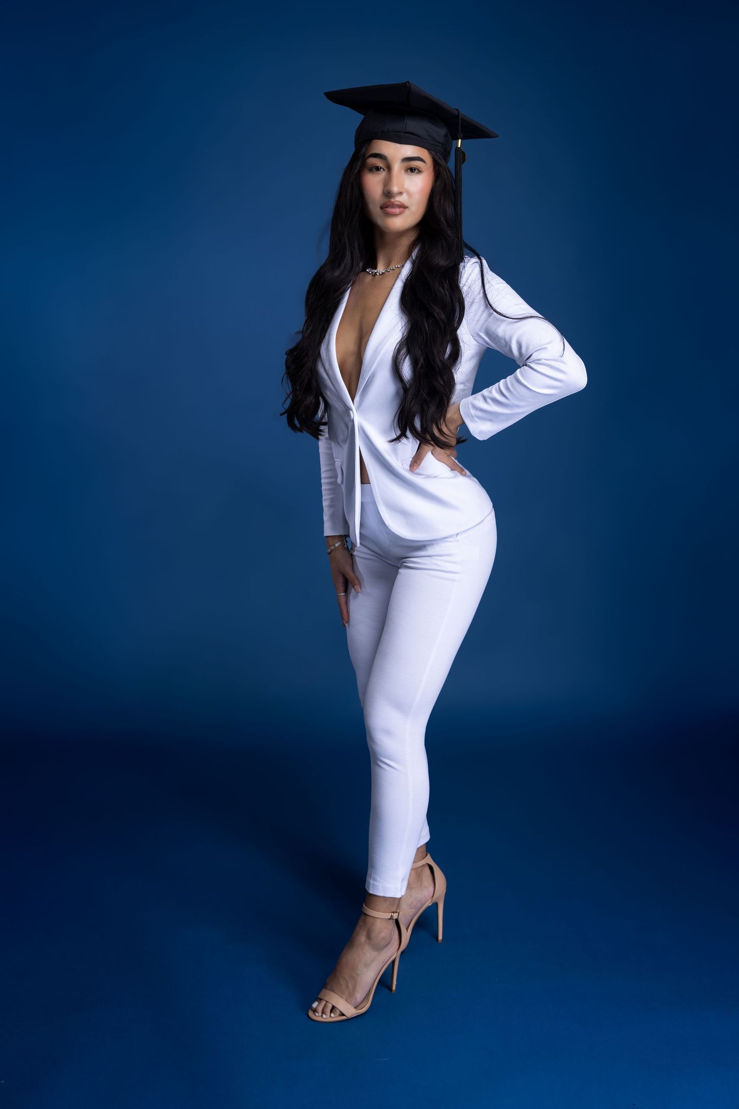 Elegant graduation photo in all-white outfit and cap against blue backdrop. Portrava Creative Studios