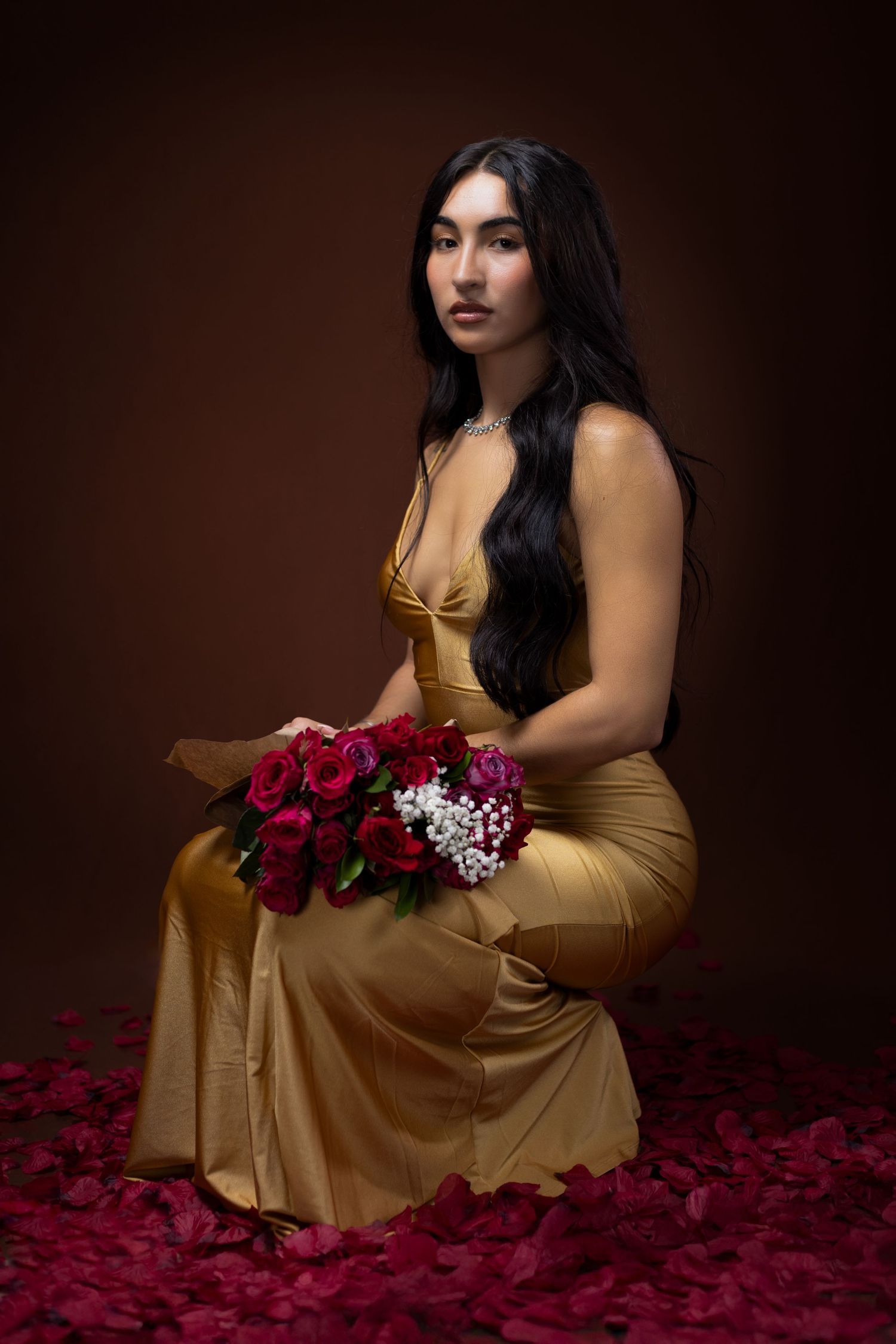 Elegant model in gold dress poses with red roses on scattered rose petals. Portrava Creative Studios