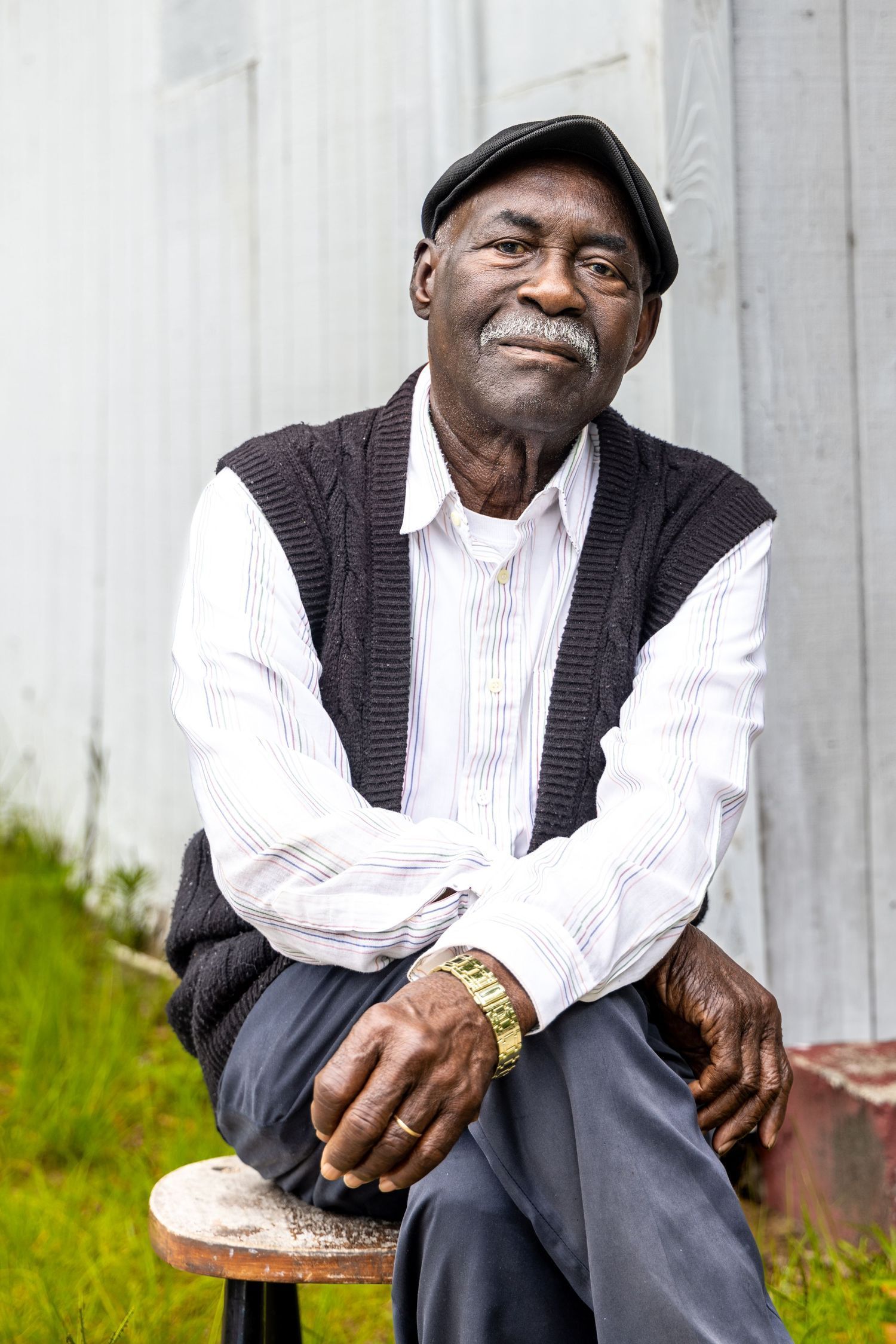 Senior person in sweater vest and cap sits outdoors with gentle smile. Portrava Creative Studios