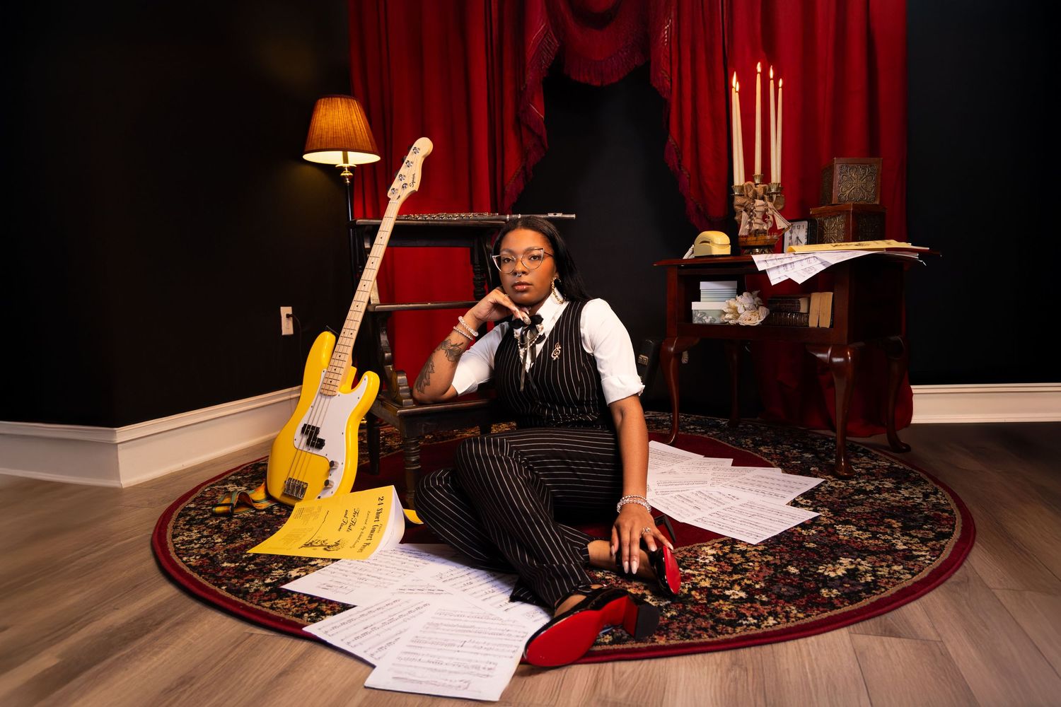 A musician sits on a round rug with a guitar and sheet music in an elegant room with red curtains. Portrava Creative Studios