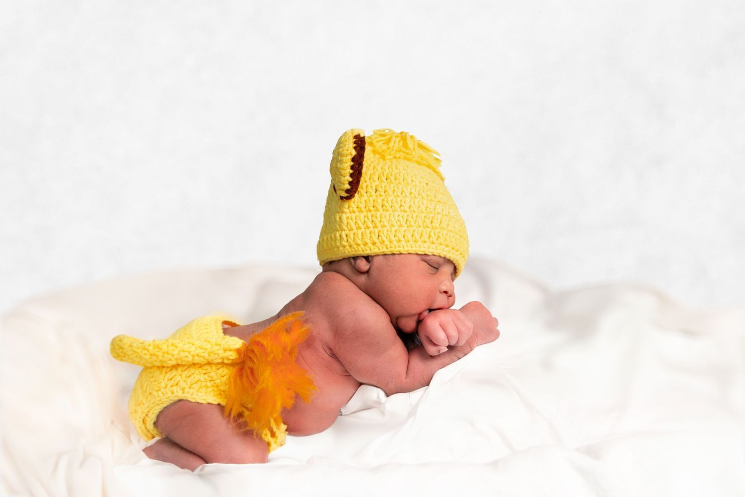 A sleeping newborn wearing a yellow knit duck outfit on white bedding. Portrava Creative Studios