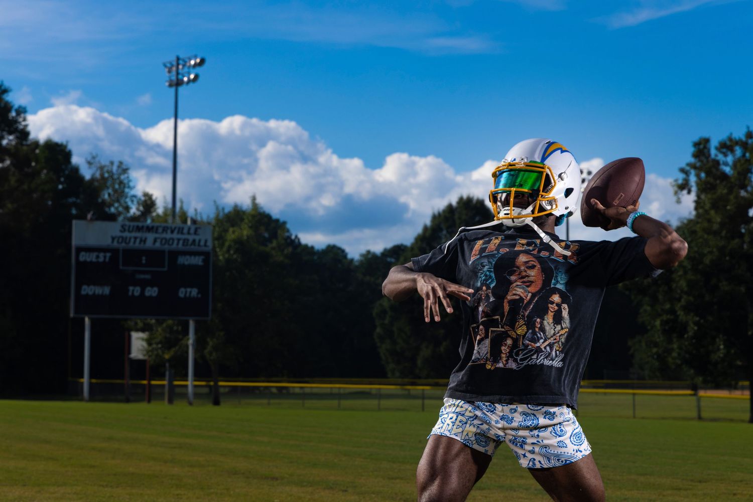 Football players practice catching drills on a sunny day at an outdoor sports field with goal posts visible.