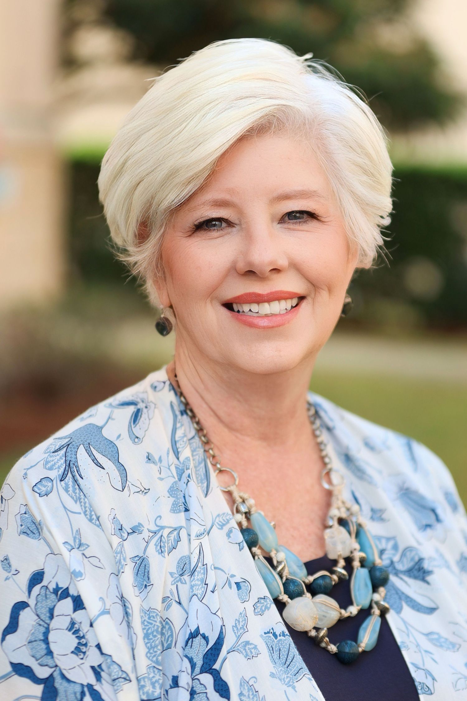 Professional headshot of a mature woman with short silver hair wearing a blue floral blouse and statement necklace. Portrava Creative Studio