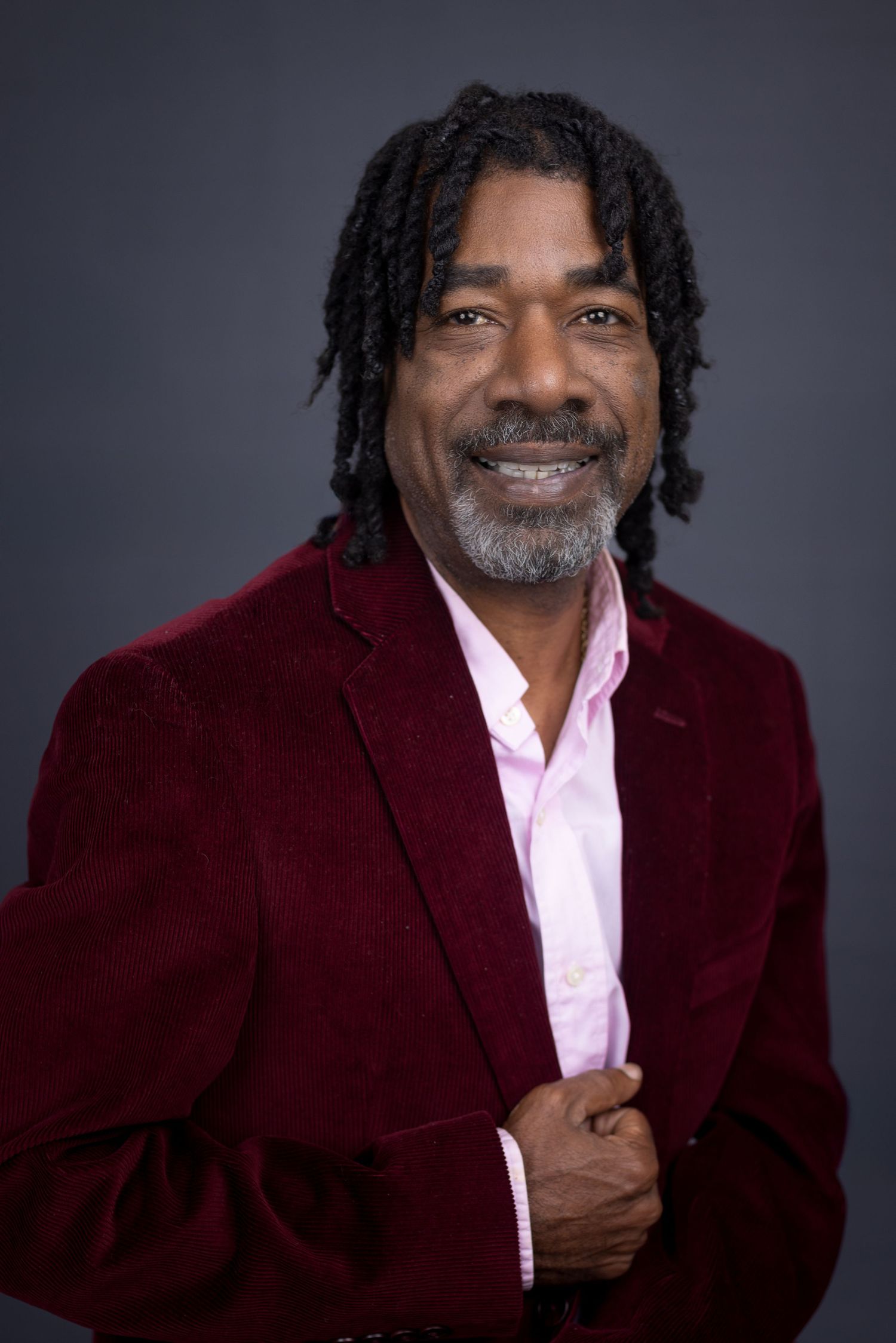 Professional headshot of a man with dreadlocks wearing a burgundy blazer and pink shirt. Portrava Creative Studios