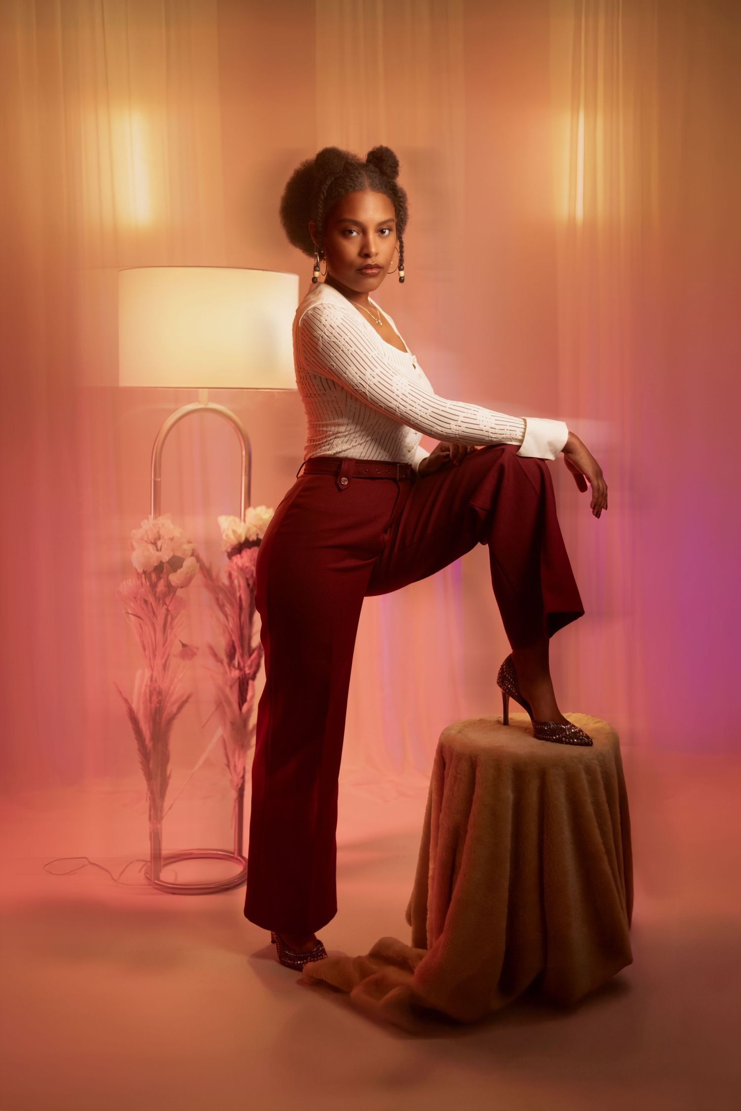 Stylish portrait with white top and burgundy pants against warm pink backdrop with table and lamp decor. Portrava Creative Studios