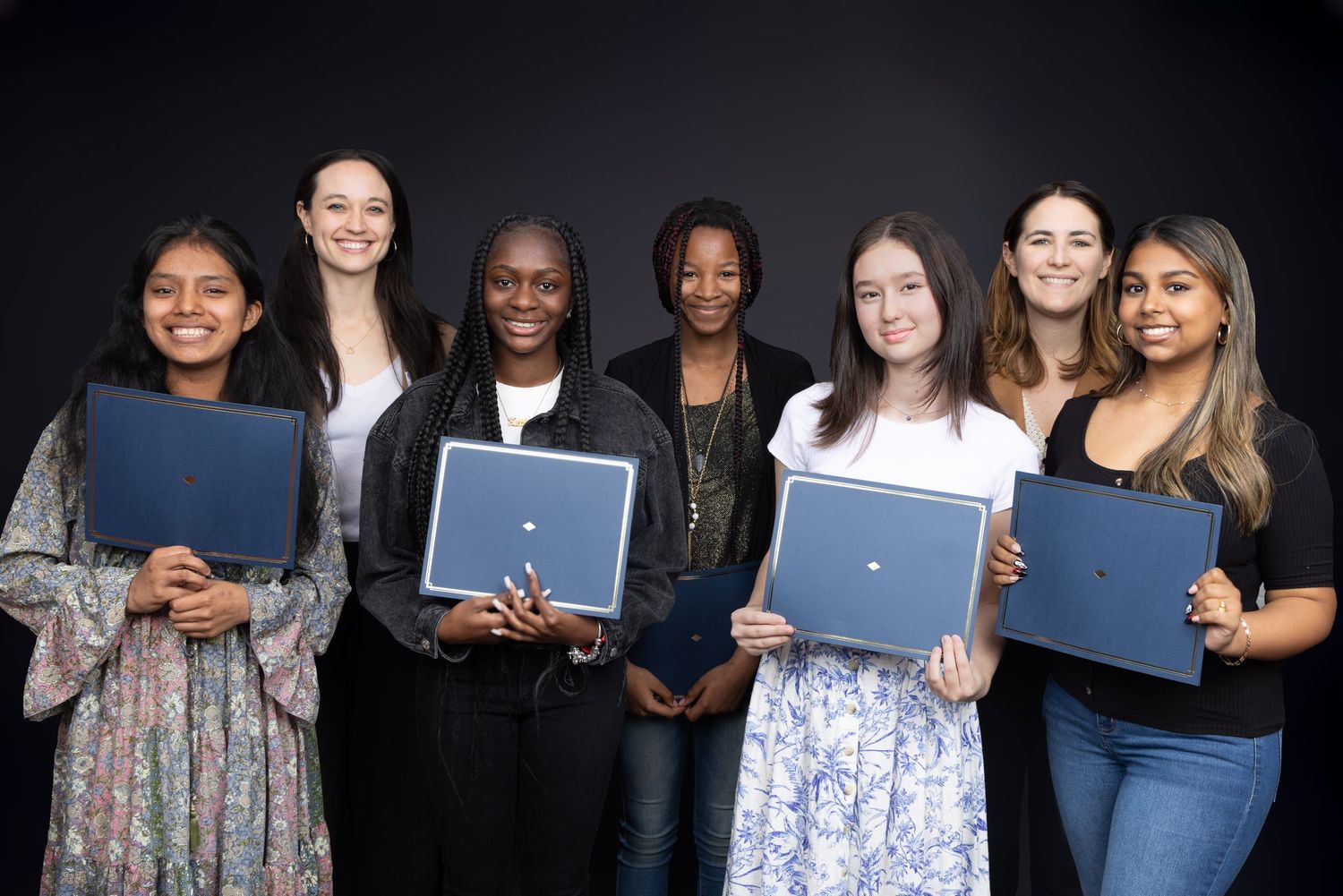 Group of graduates holding blue diploma folders pose together for formal photo. Portrava Creative Studios